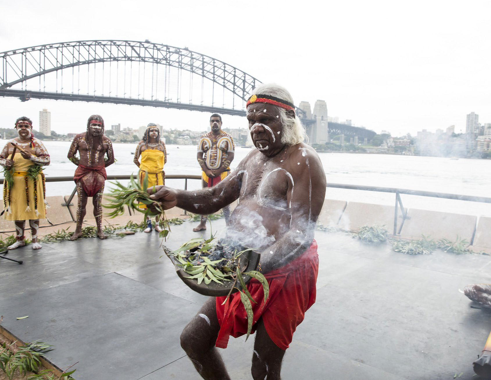 An Indigenous Australian man, with traditional face and body paint, holding a bowl of leaves and performing a cultural ritual on a stage near a river, with a bridge and city skyline in the background.