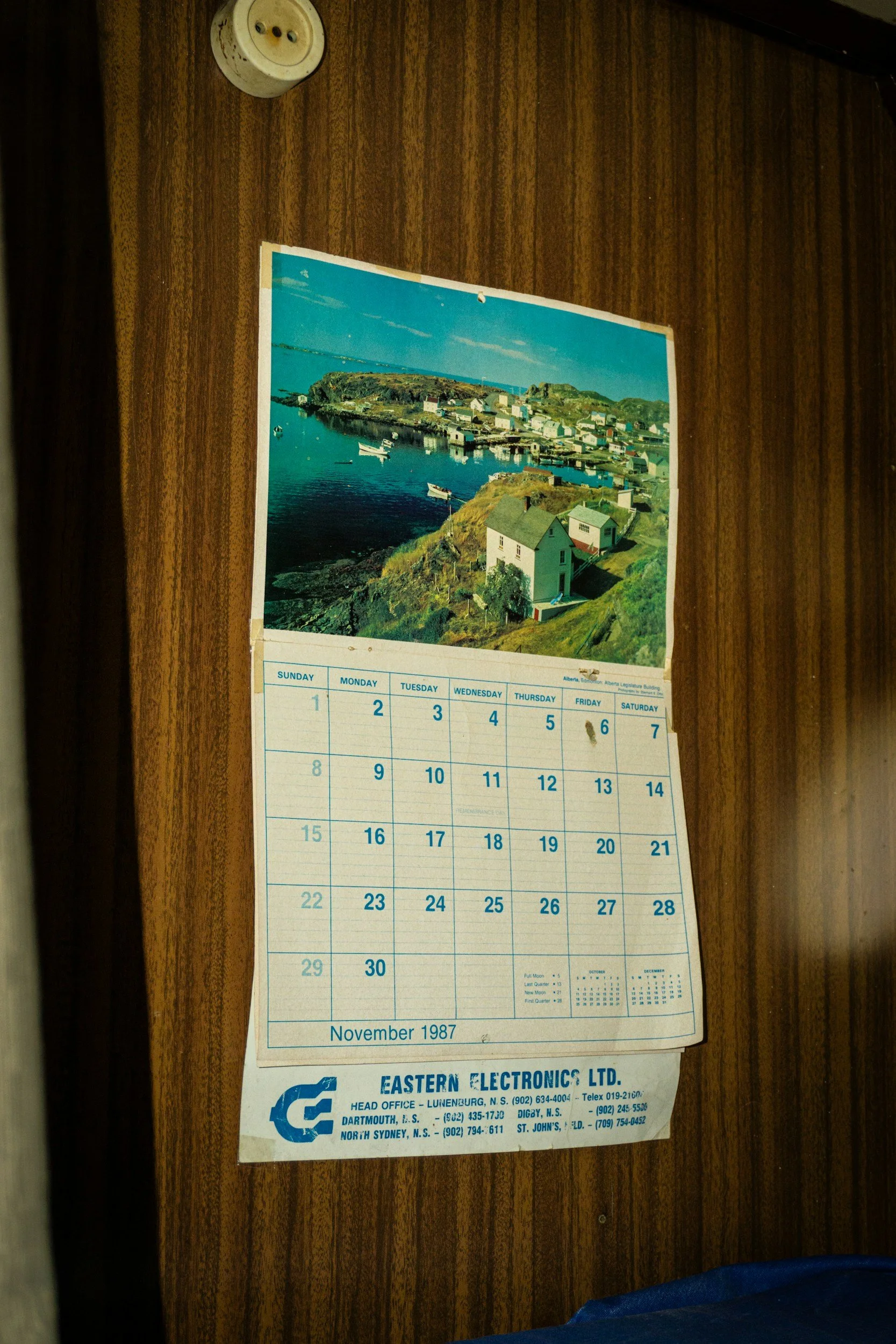 A calendar hanging on a wooden wall, showing November 1987, with a scenic photograph of a coastal village at the top.