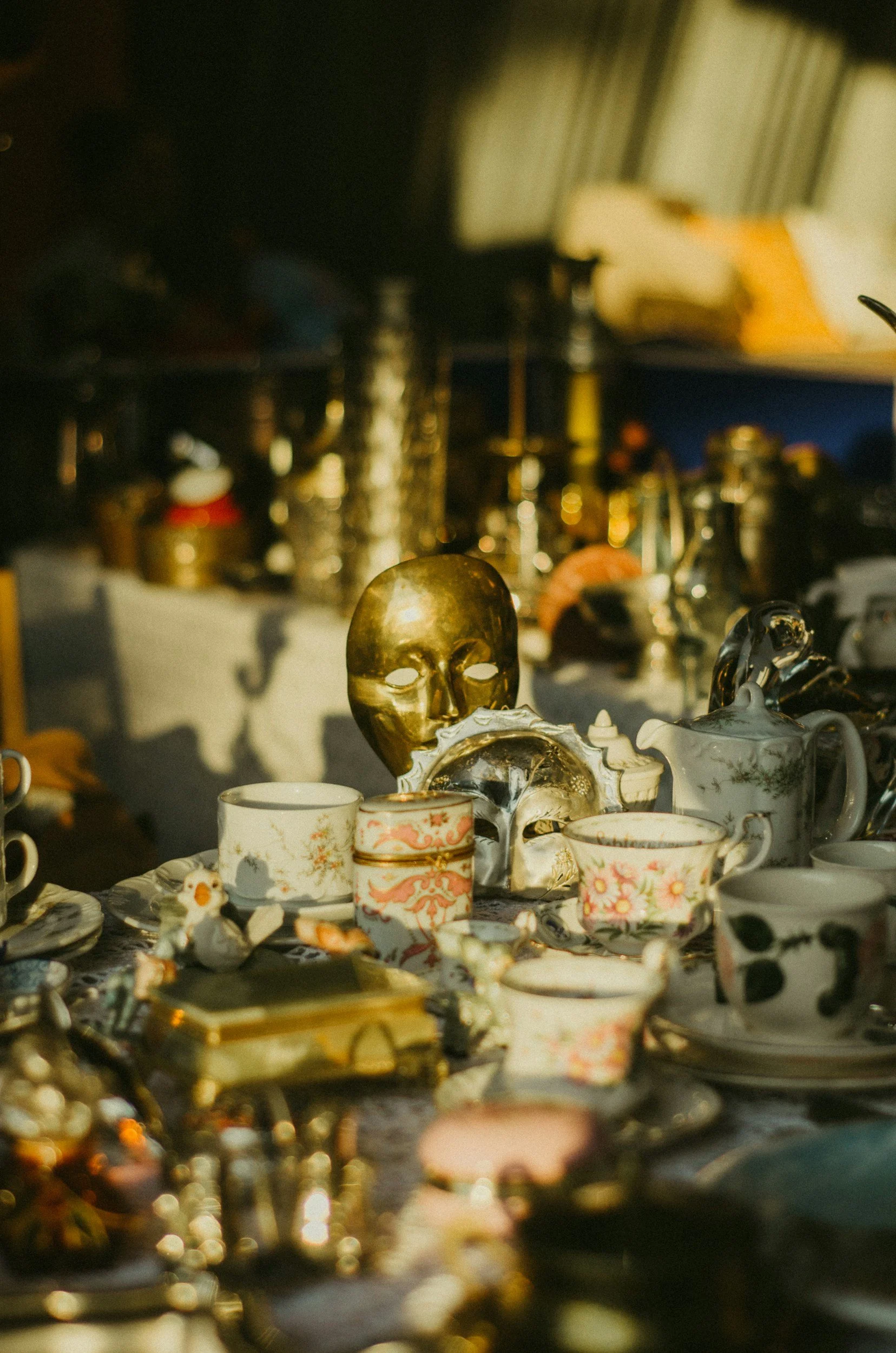 Various vintage and decorative objects including a gold-colored skull, teapots, cups, and small figurines displayed on a surface at a flea market or antique shop.