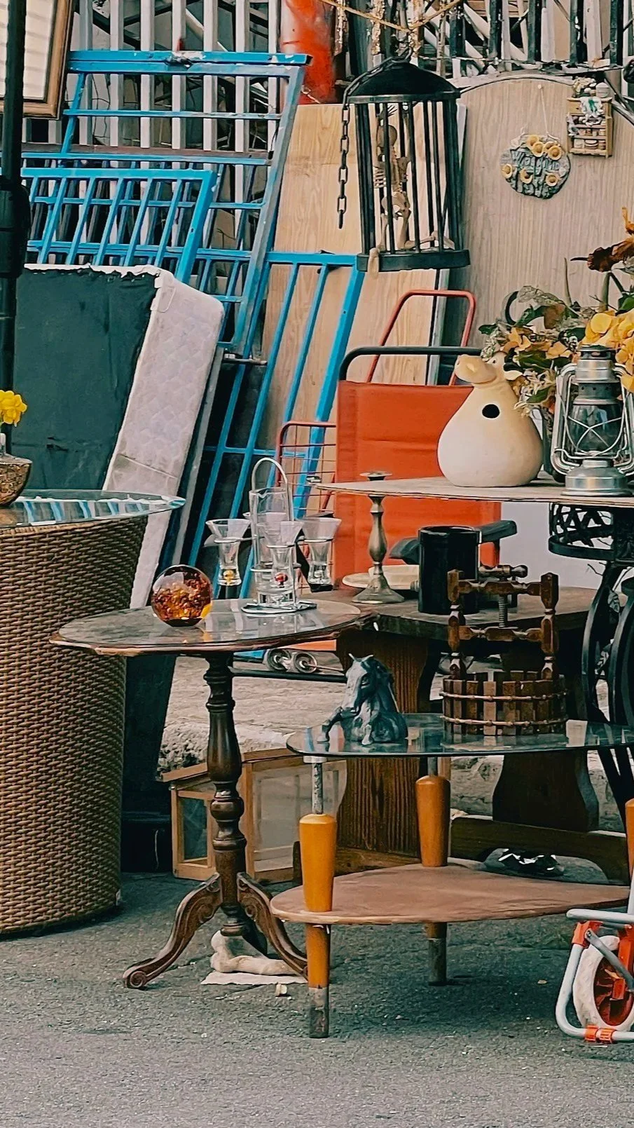 An outdoor scene with various household items and furniture, including tables, chairs, lamps, and decorative objects, arranged in a cluttered display.