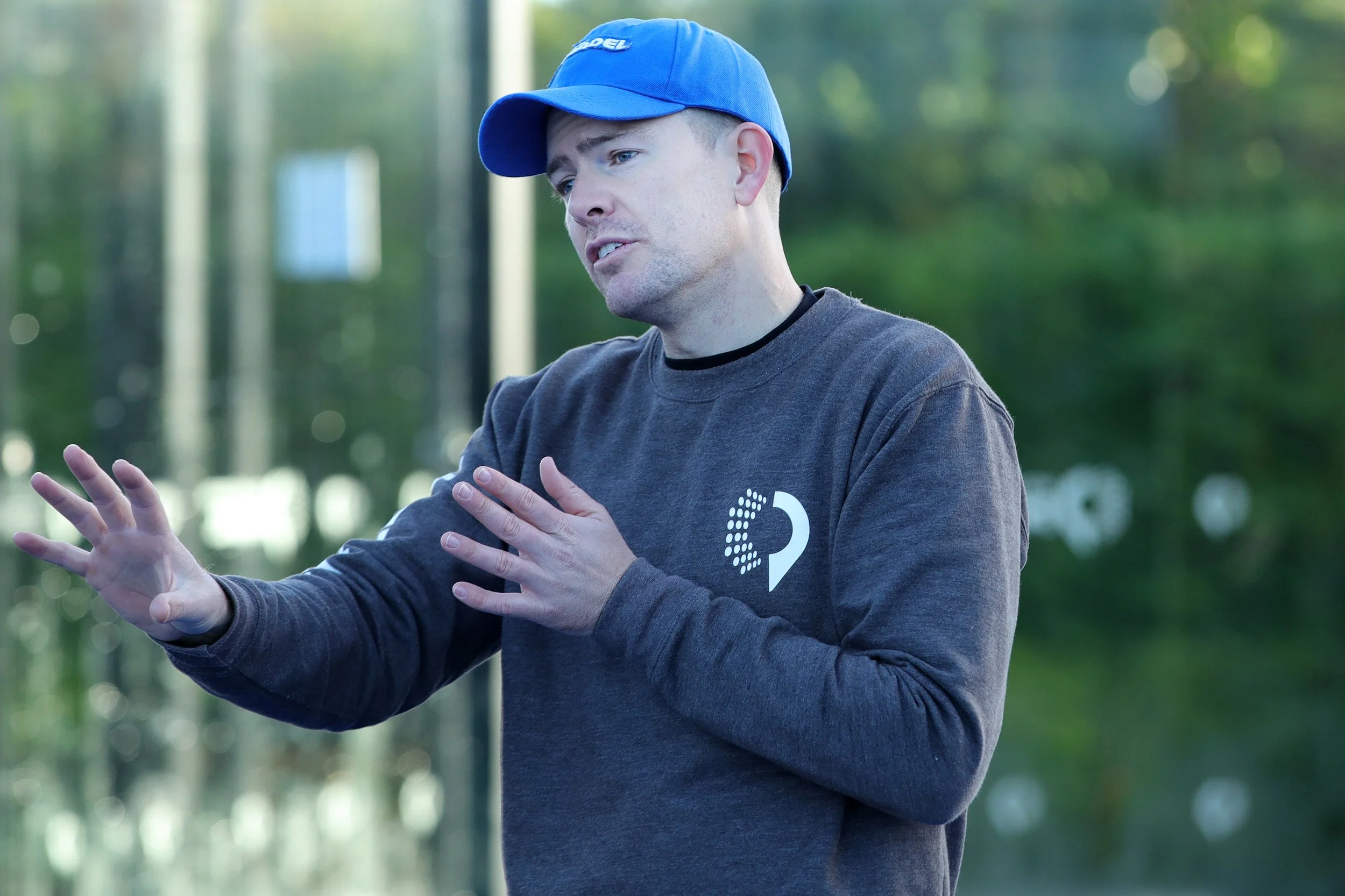 A young man wearing a blue baseball cap and a dark gray sweatshirt with a logo on it. He appears to be outdoors in a park, gesturing with his hands while looking serious. A padel coach.