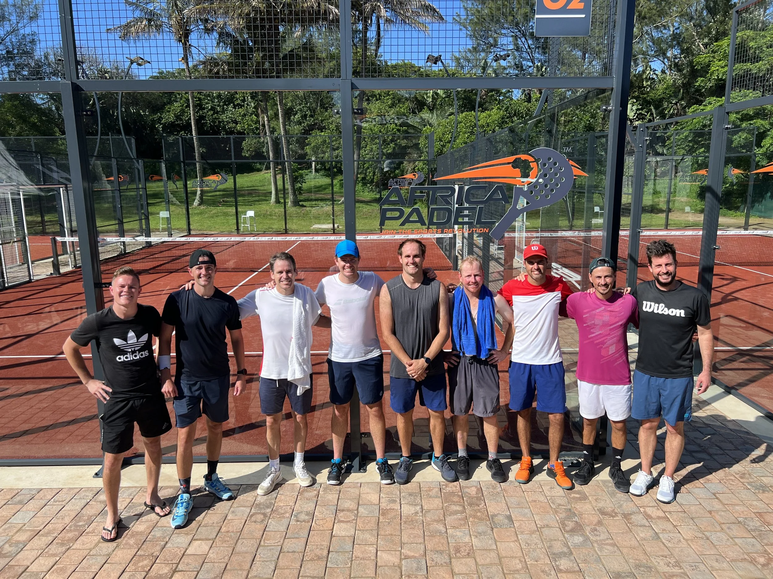 A group of eight men standing on a padel court, smiling at the camera, with a padel court fence, lush green trees, and blue sky in the background. There are signs for 'Africa Padel' on the fence.