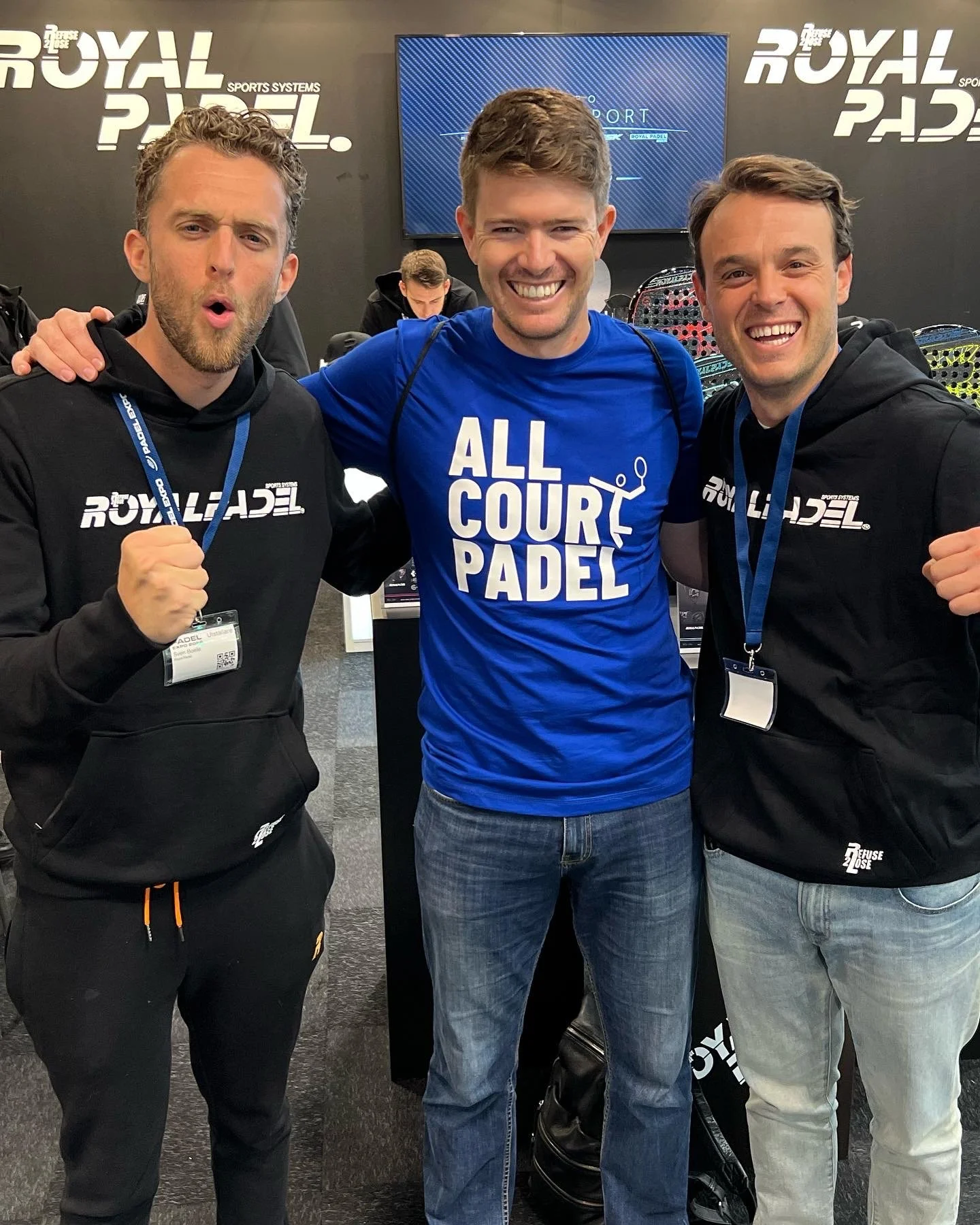 Three men posing at a padel event, wearing casual clothing with event branding. They are smiling and showing enthusiasm.