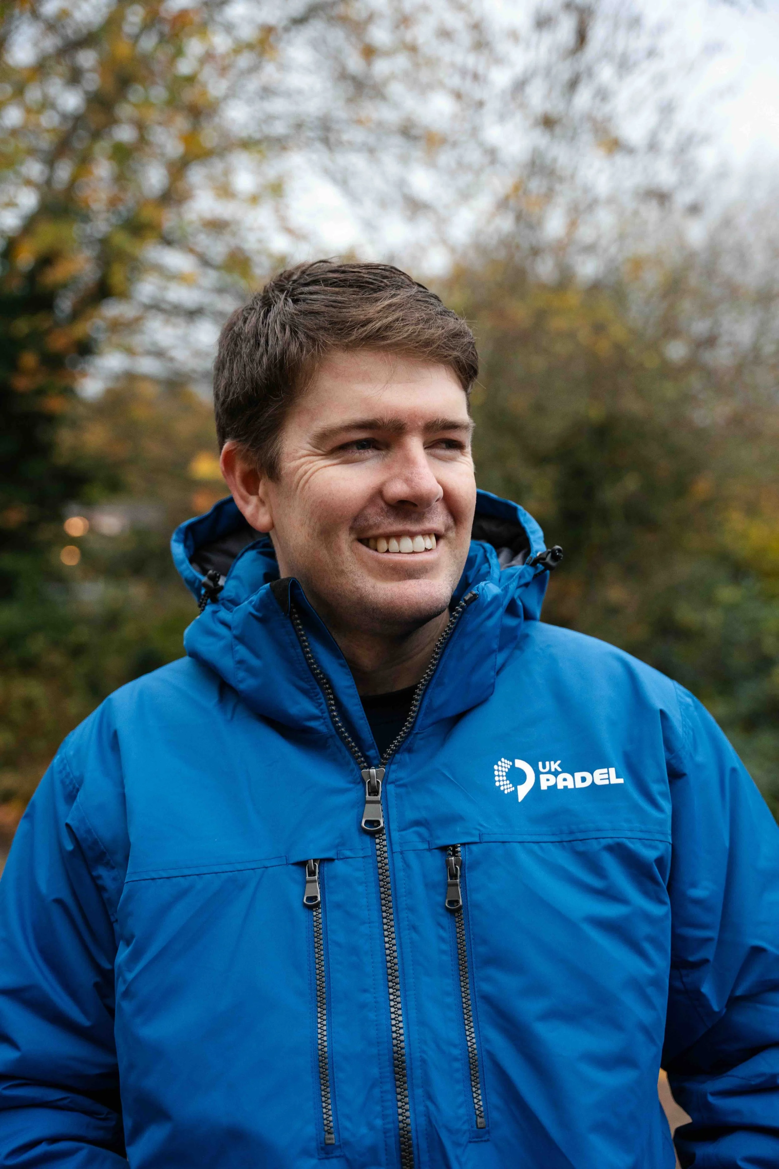 A smiling man in a blue jacket standing outdoors with autumn trees in the background.