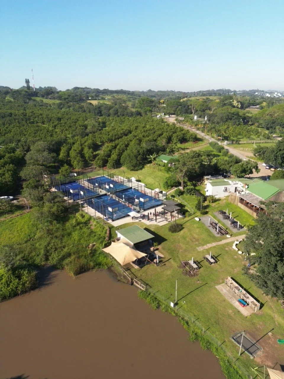 Aerial view of padel courts, a pond, and green landscape with trees and buildings in the distance under a clear blue sky.