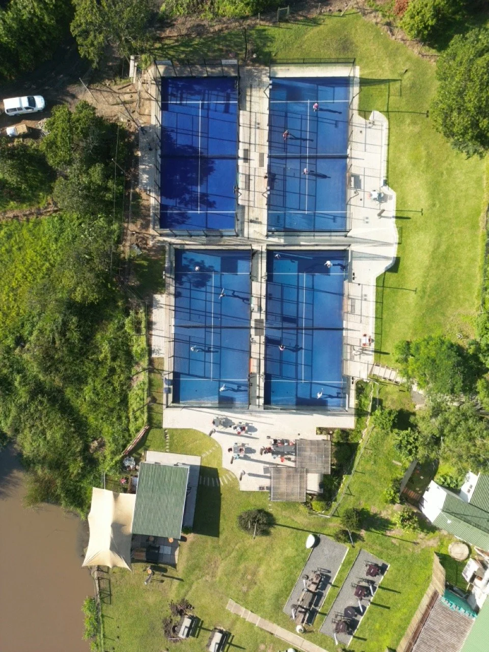 Aerial view of four padel courts with players, surrounded by green trees and grassy areas, and small buildings nearby.
