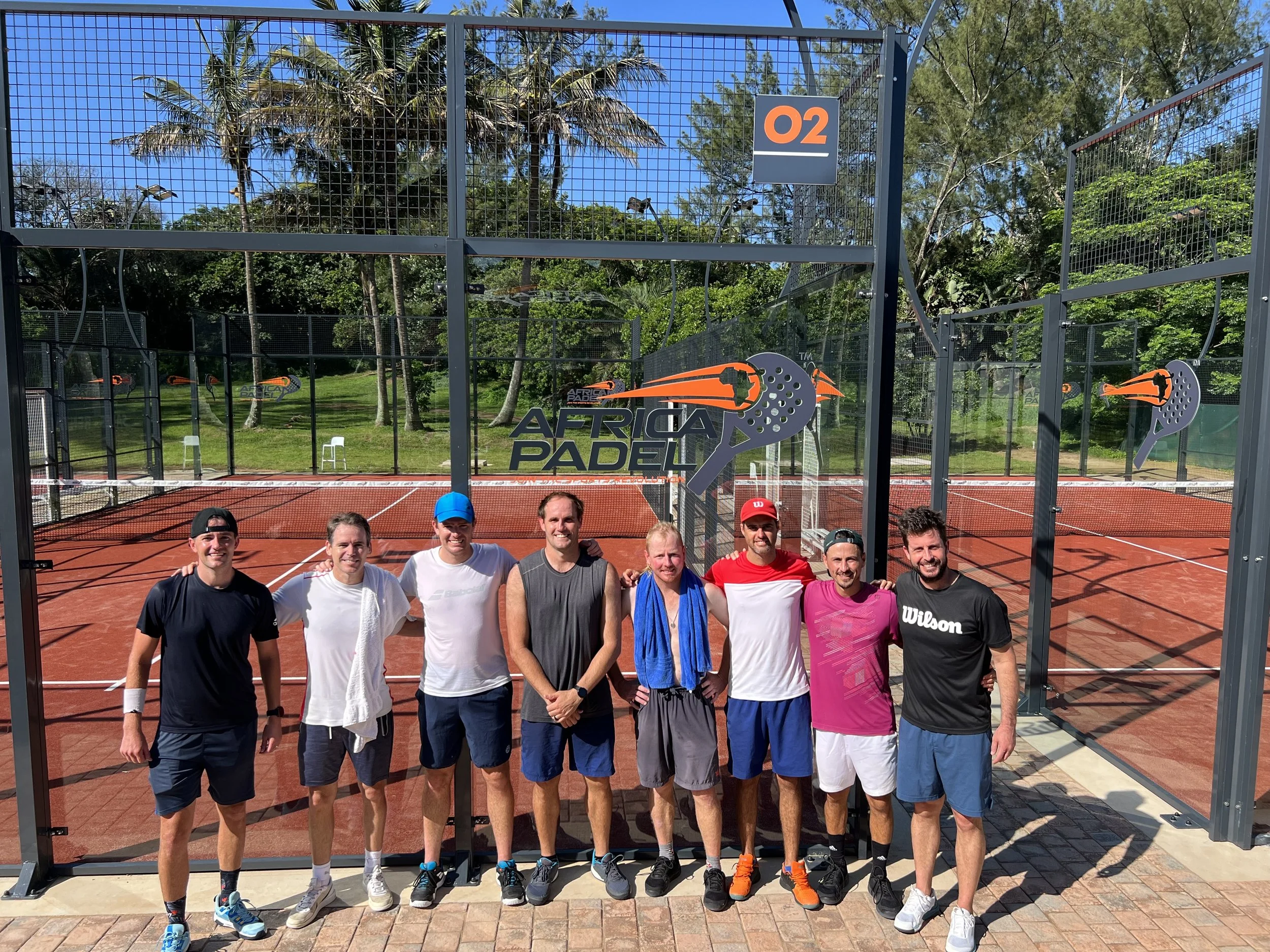 Group of men standing together on a padel court, posing for a photo, with palm trees and lush greenery in the background, sunny weather.