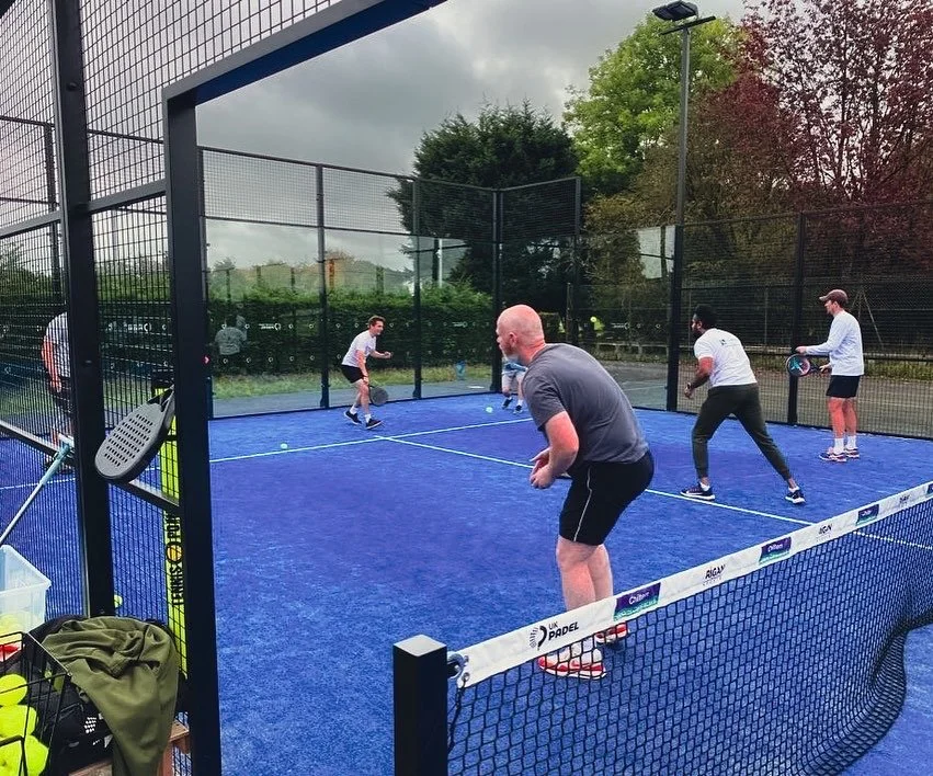 Five people playing padel on a blue court surrounded by a tall black fence, with trees and a cloudy sky in the background.
