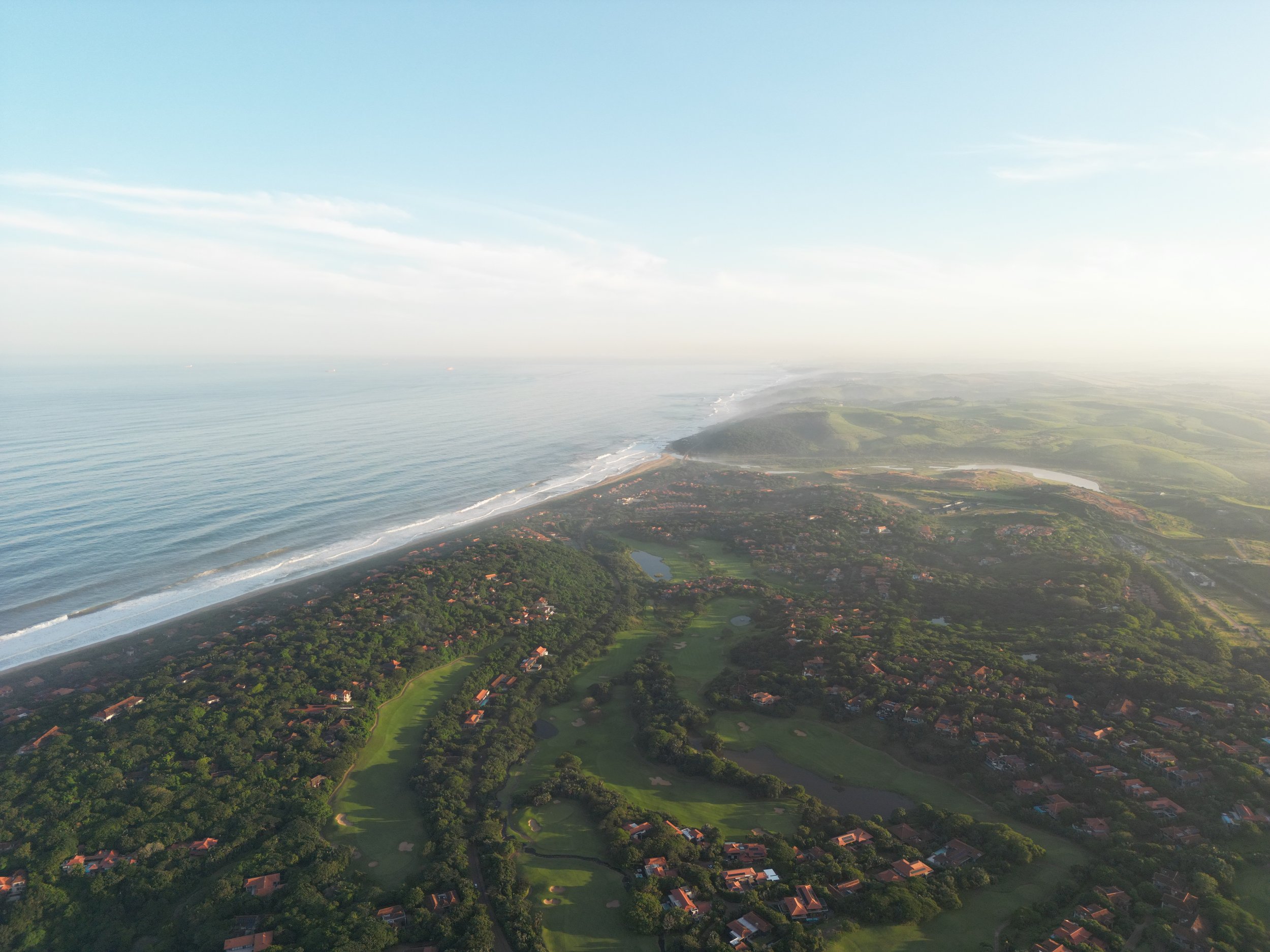 An aerial view of a coastline with the ocean on the left and green land with houses and a golf course on the right.
