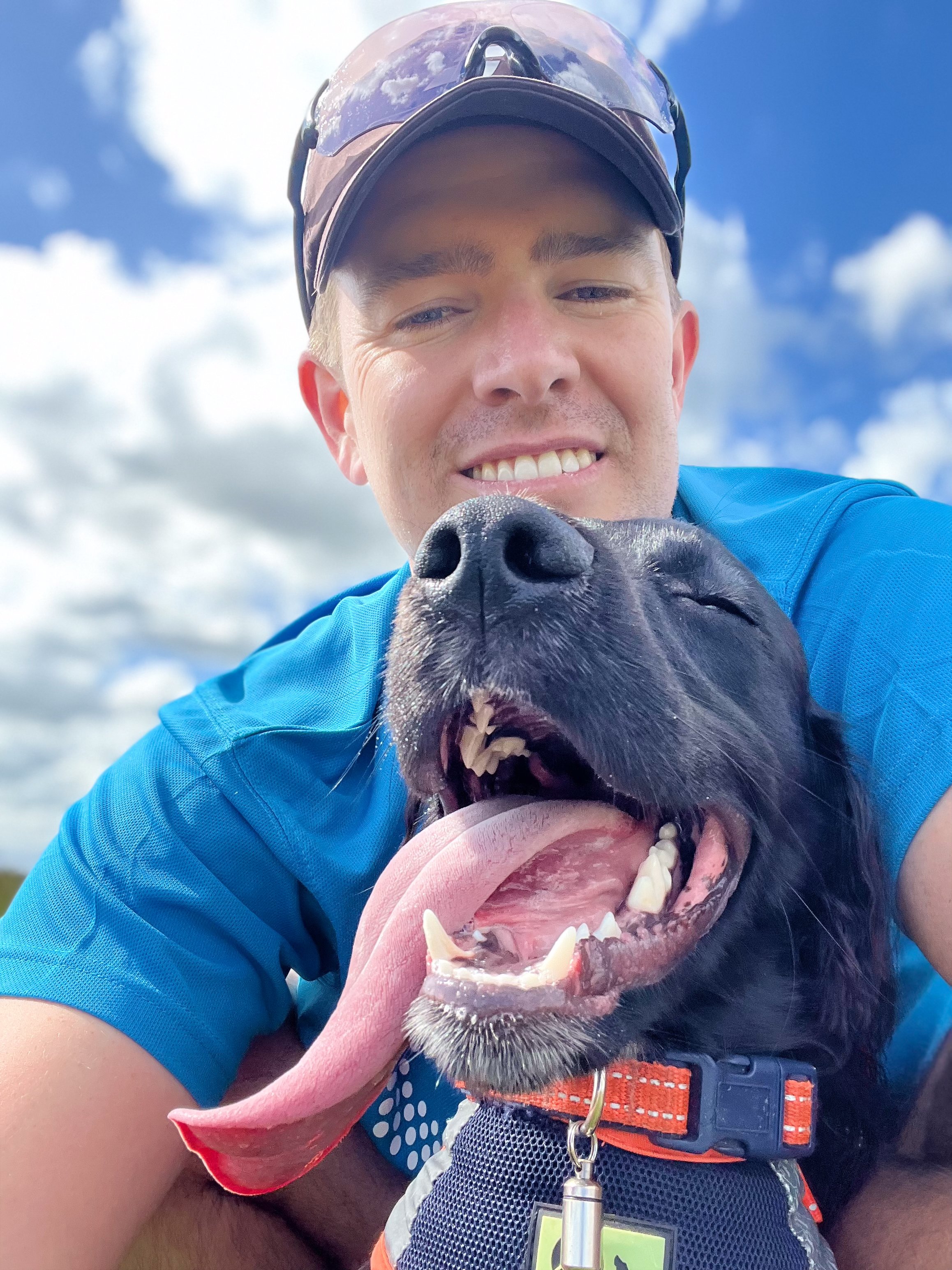 A person wearing a cap and sunglasses taking a selfie with a black dog, both outdoors under a blue sky with clouds. The dog appears excited, with its tongue out and mouth open.