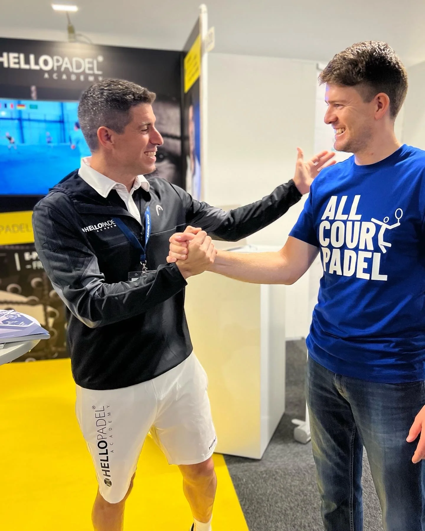 Two men smiling and shaking hands in a sports setting. One wearing a black jacket with 'HELLOPADEL ACADEMY' and white shorts, the other in a blue t-shirt with 'ALL COURT PADEL' and jeans.