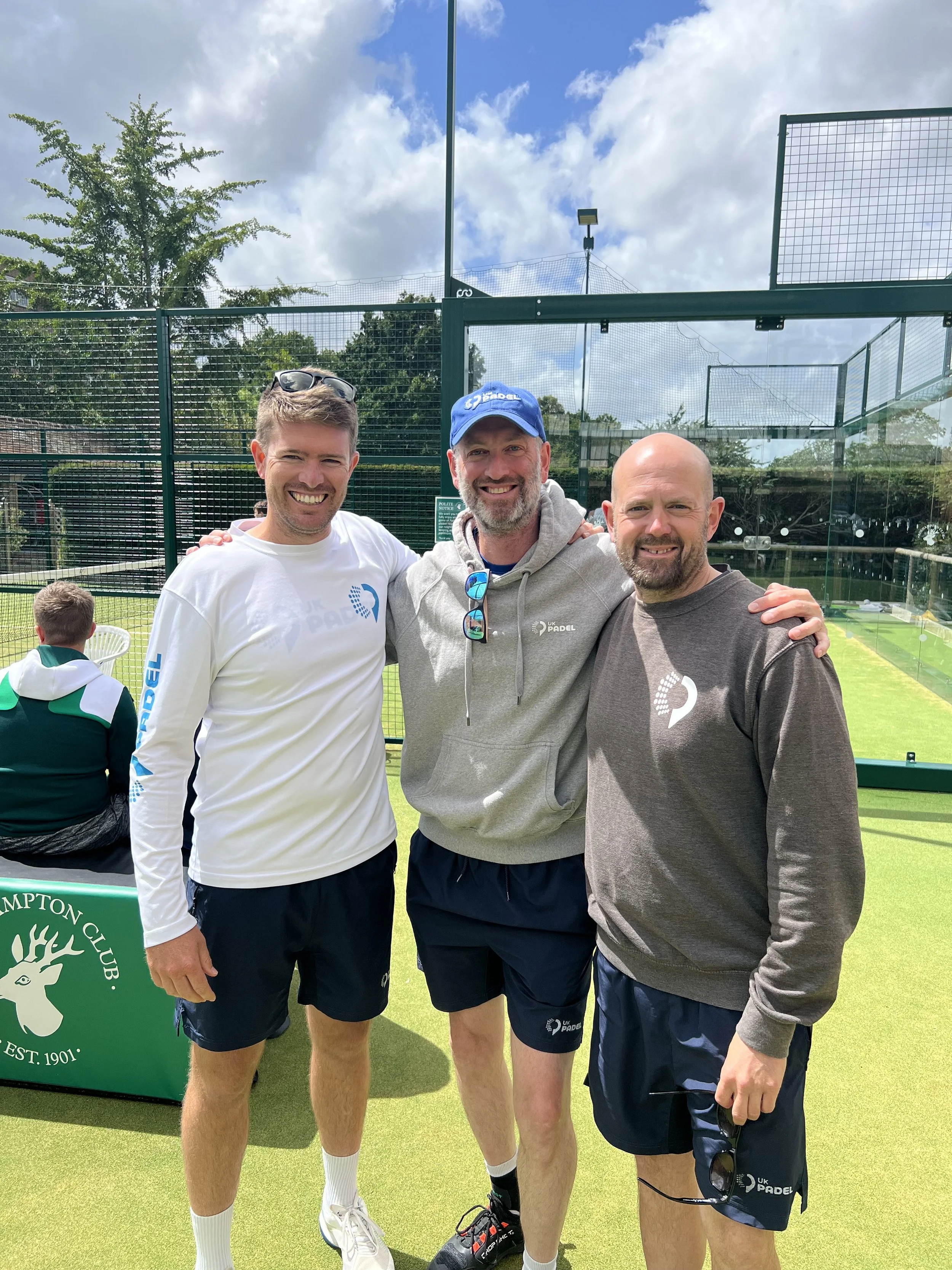 Three men standing together on a green padel court, smiling, with tennis nets in the background. They are dressed in athletic clothing and are outdoors on a sunny day with a partly cloudy sky.