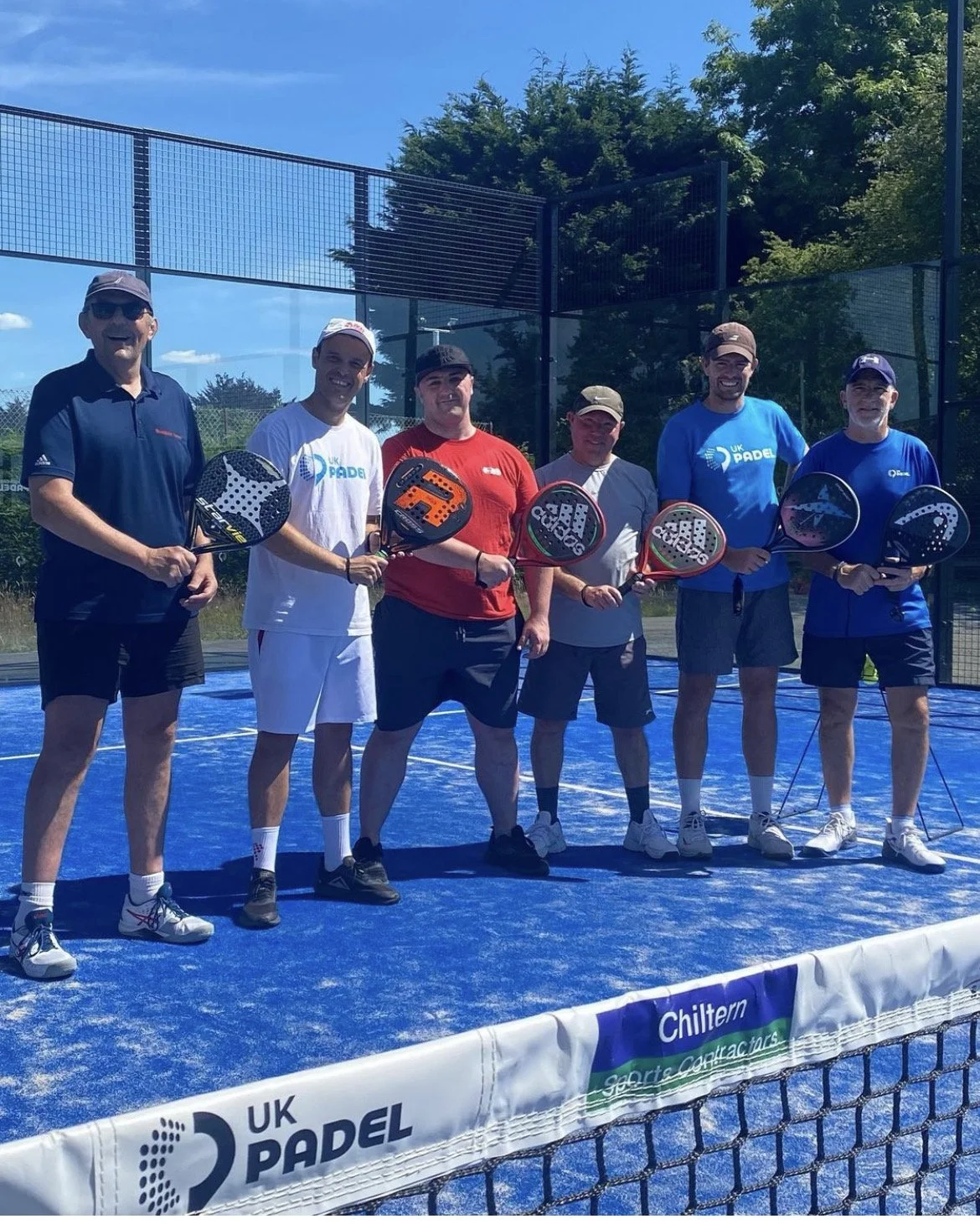 Group of six men standing on a blue padel tennis court holding paddles, smiling at the camera. They are dressed in sportswear and caps, with a tennis net and a fence in the background under a clear blue sky.