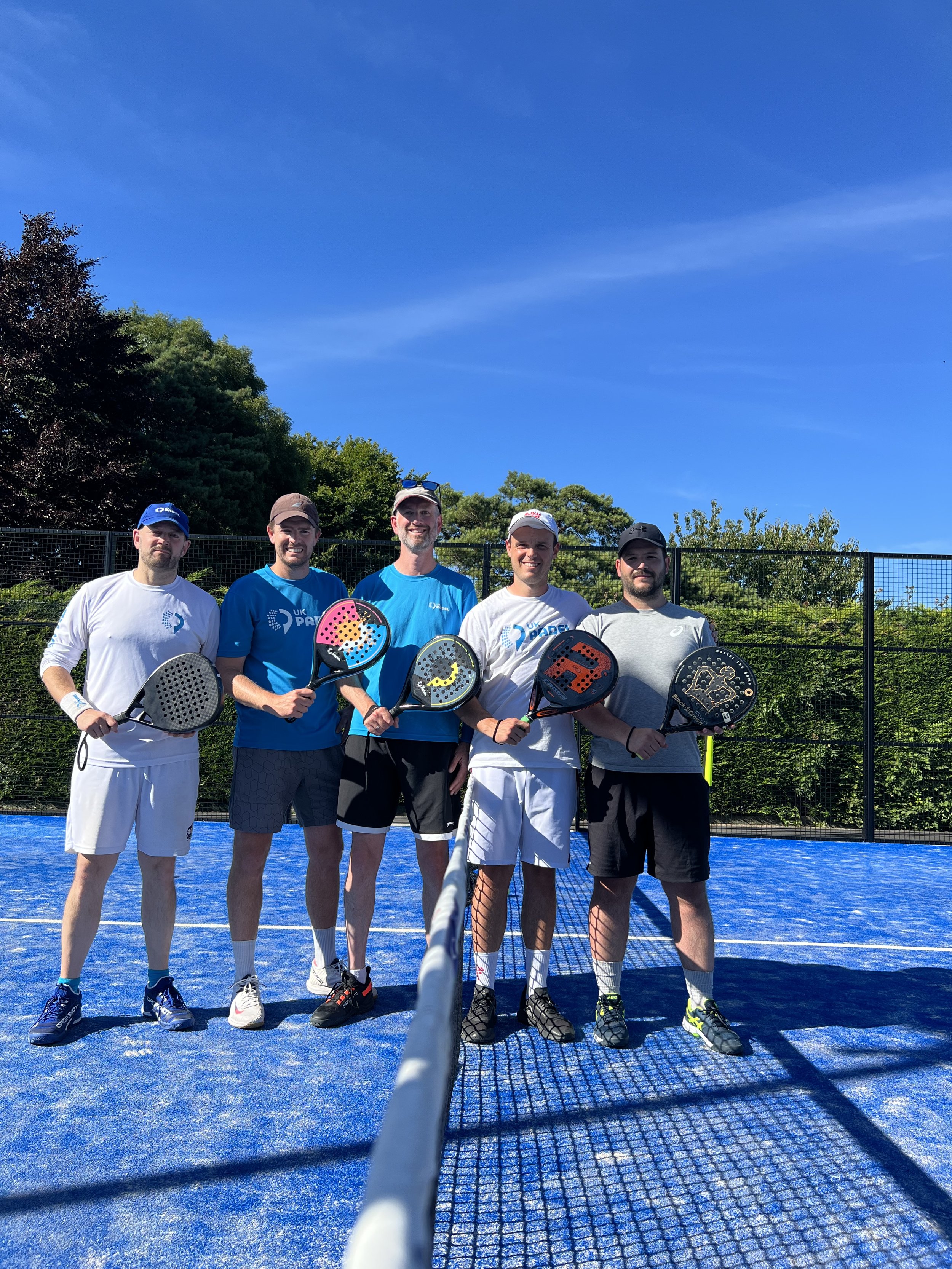 Five men standing on a blue pickleball court holding paddles, with trees and a black fence in the background, under a clear blue sky.