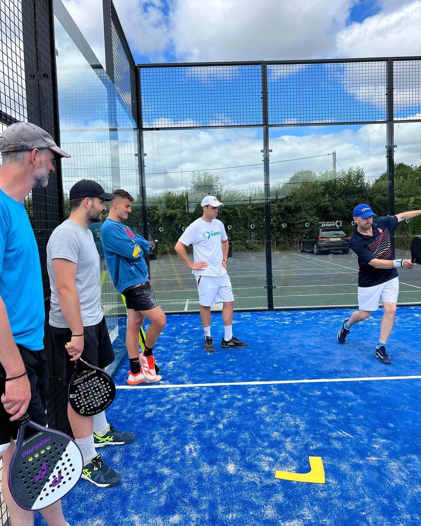 Group of men practicing padel on an outdoor court with a bright blue surface and glass walls.