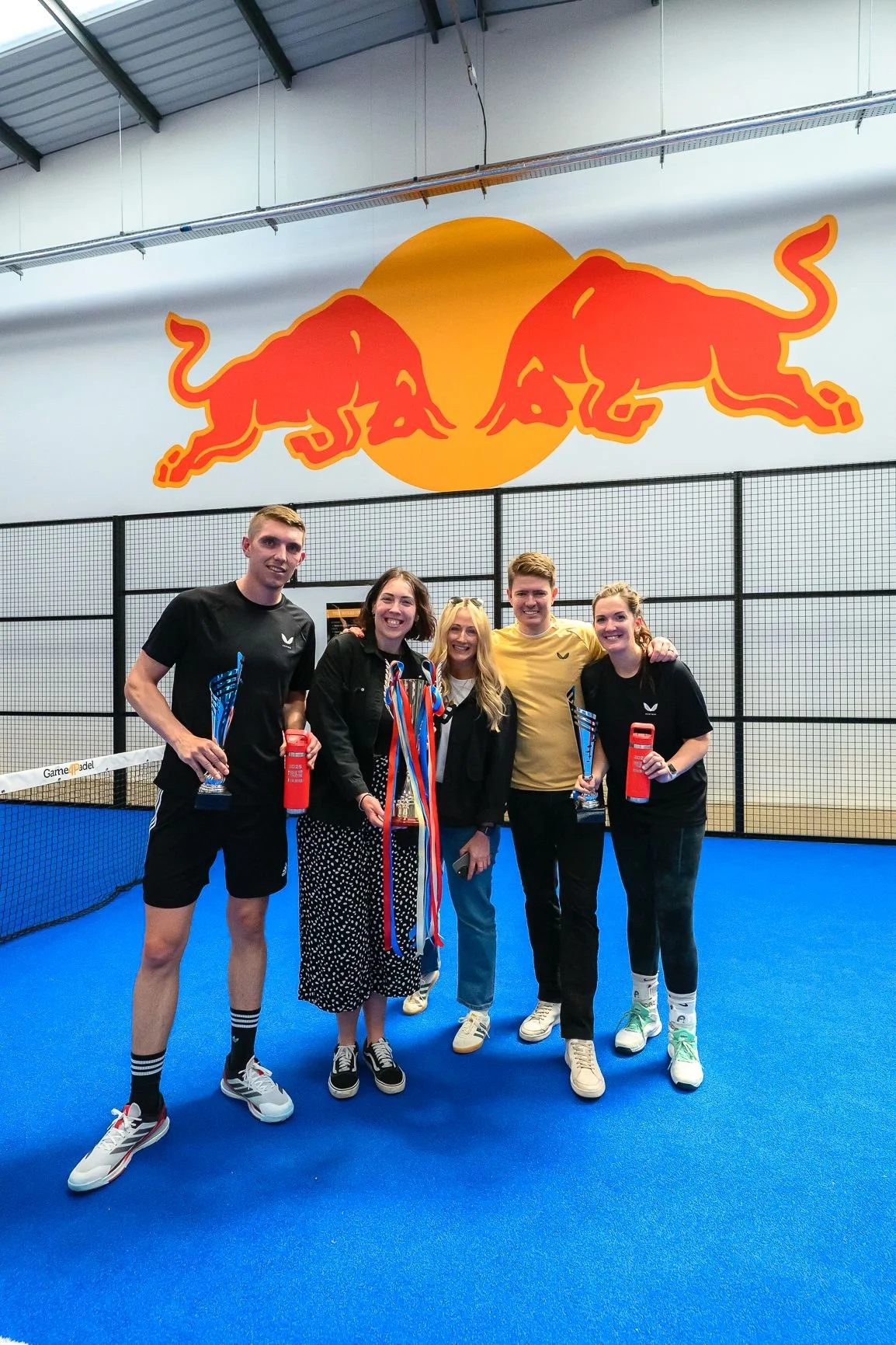 Group of five people celebrating on a padel court, holding trophies and medals with a Red Bull logo in the background.