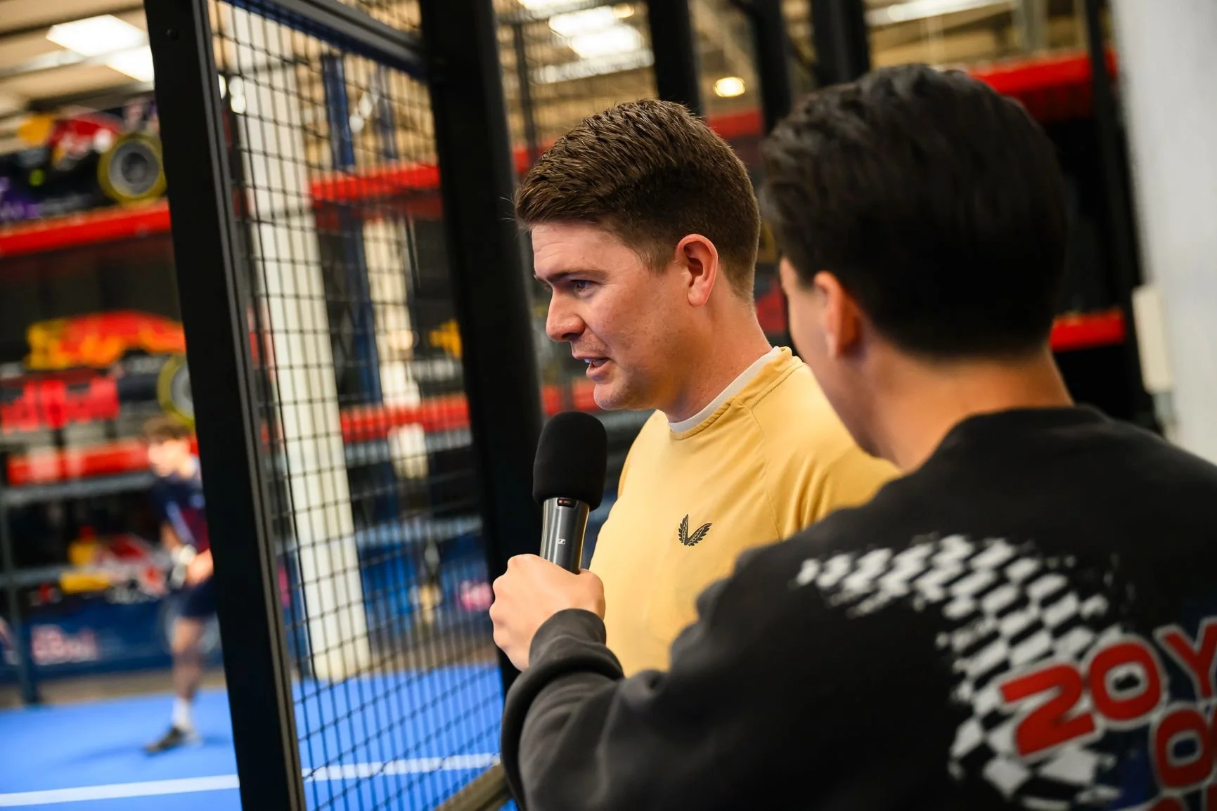 A man in a yellow shirt holding a microphone is speaking to another man in a black jacket with red and white patterned sleeves, in an indoor padel facility with shelves of F1 cars in the background.