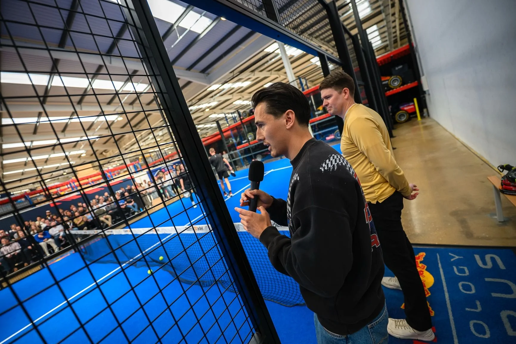 Two men watch a tennis match through at an indoor sports facility. One man holds a microphone and appears to be speaking, while the other stands behind him, both focused on the game. The blue tennis court and spectators can be seen in the background.
