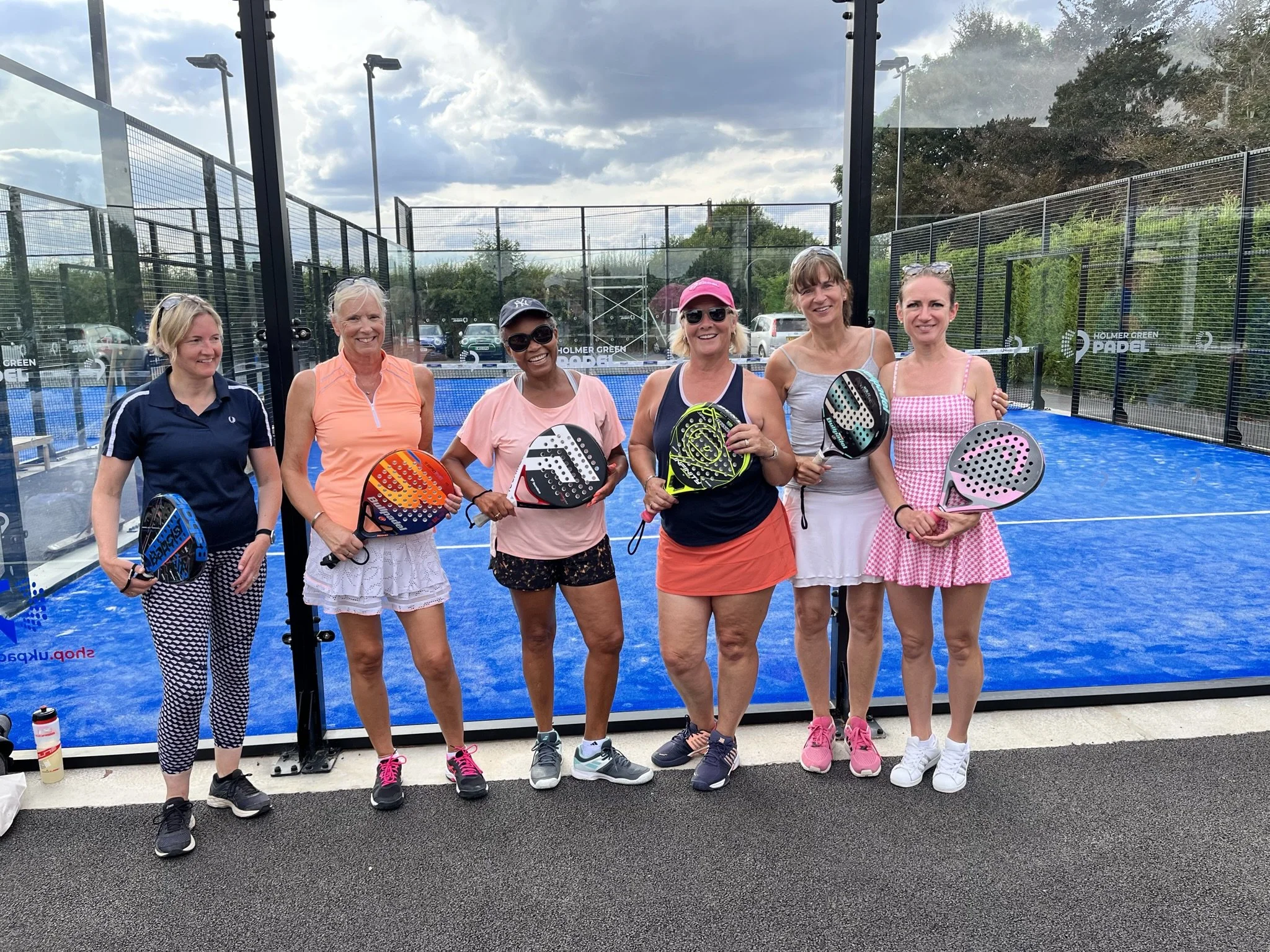 Six women standing on a padel court holding padel rackets, smiling at the camera. The court has a blue playing surface and black fencing, with trees and parked cars in the background.