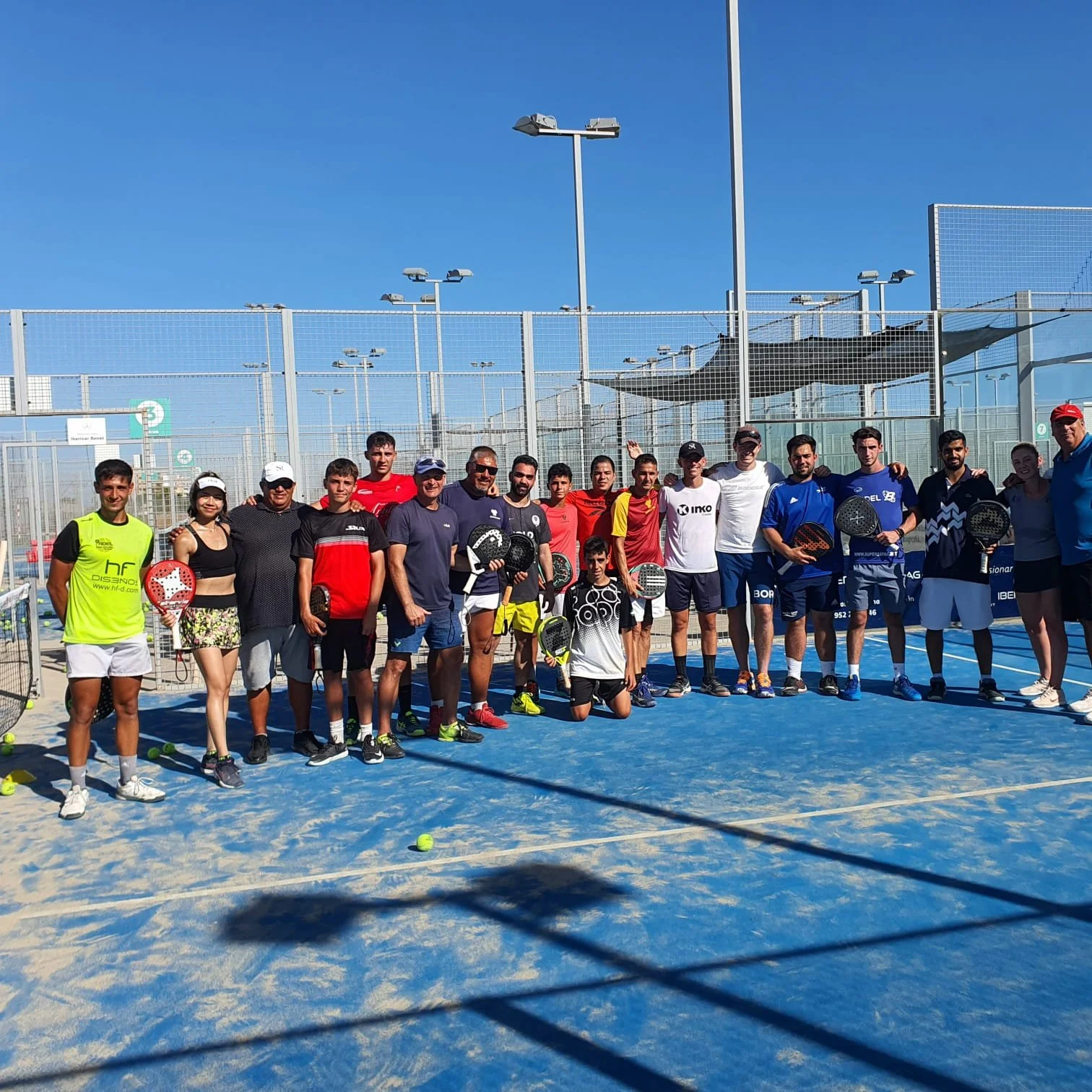 Group of people on a padel court after a game, some holding rackets, with a fence, blue sky, and tennis nets in the background.