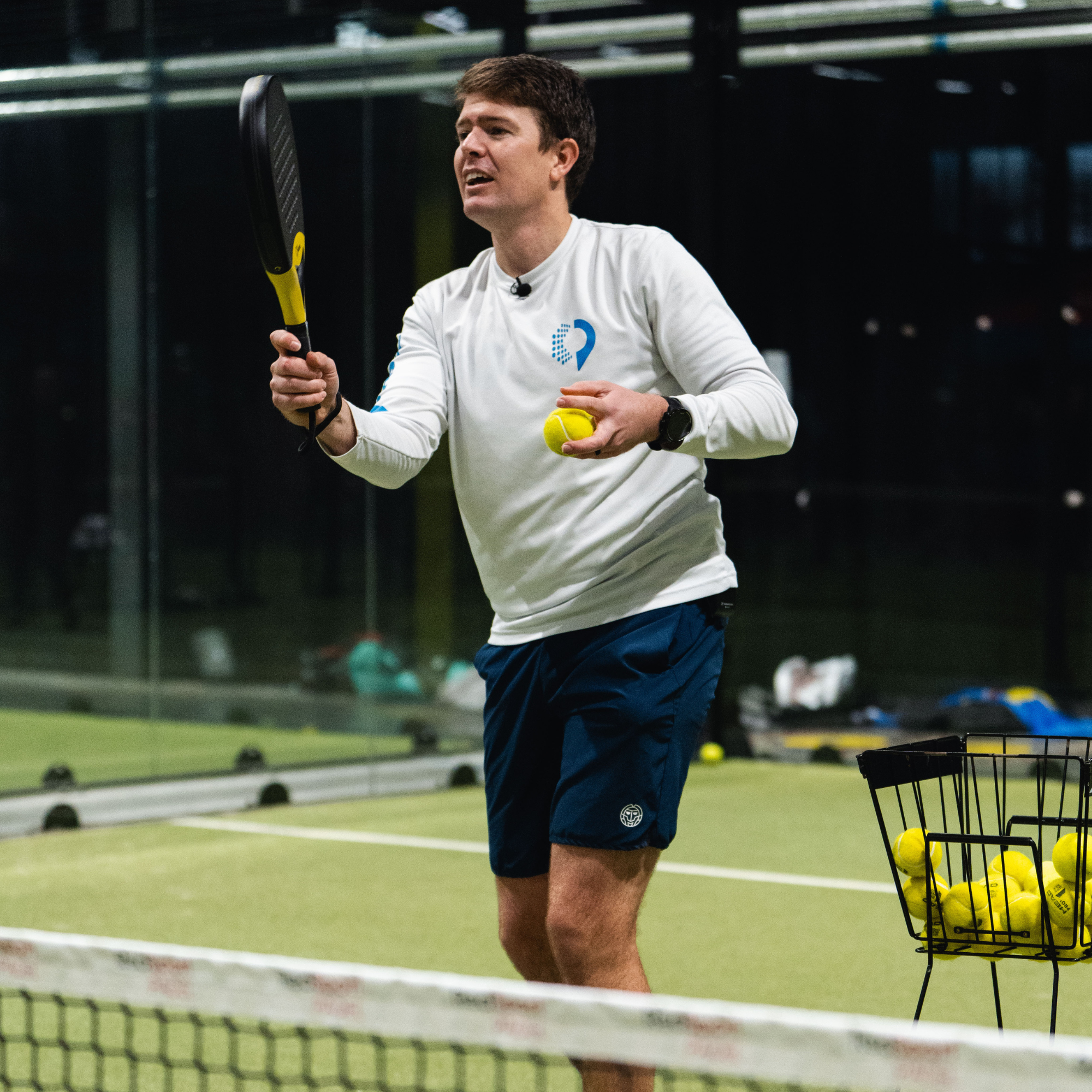 A man holding a padel racket and tennis ball in an indoor padel court.