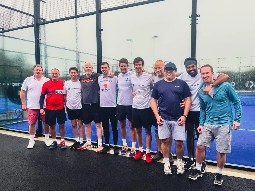A group of eleven men standing together on a padel tennis court, smiling and posing for a photo.
