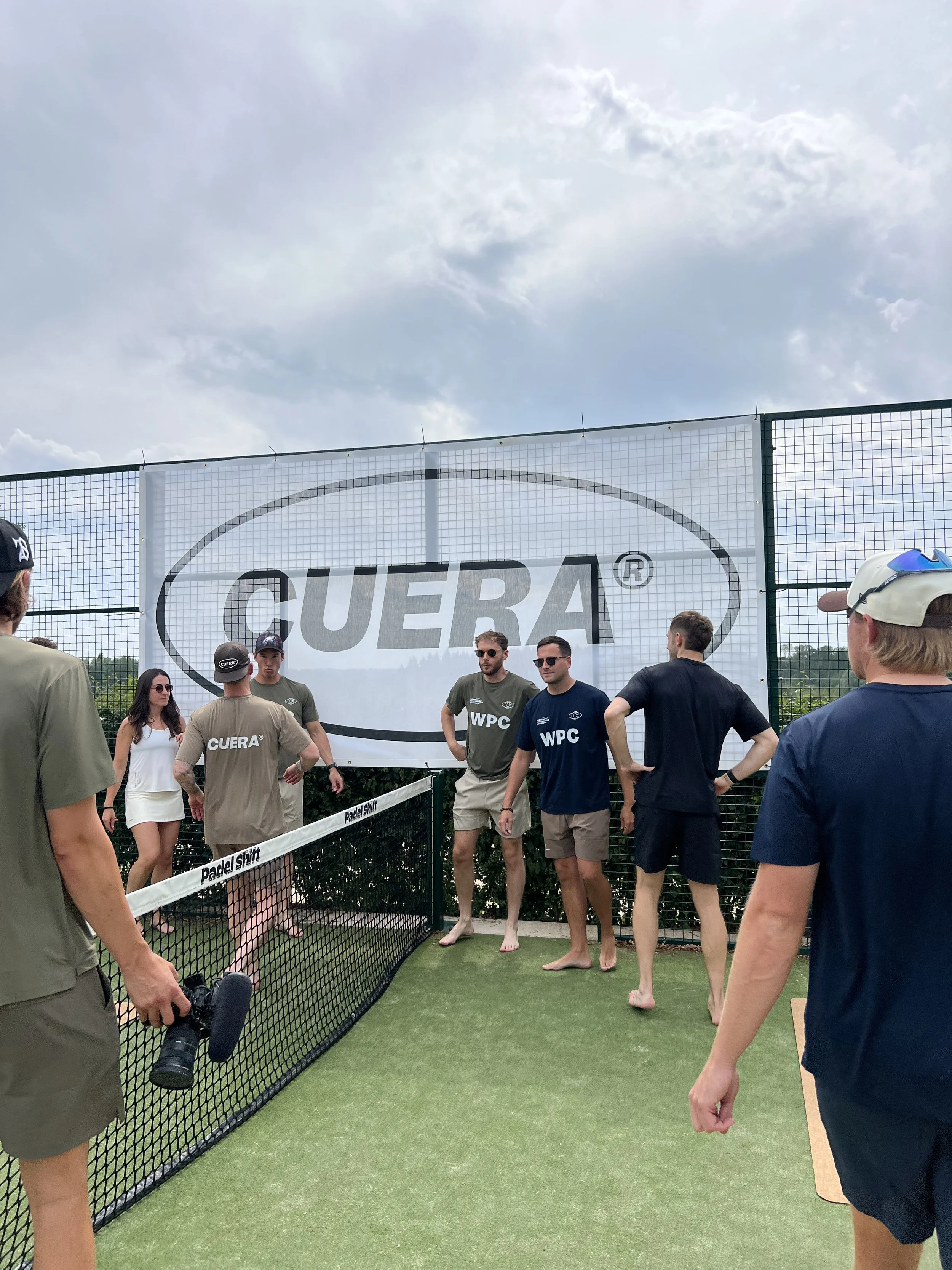 Group of people gathered near a fenced padel court during daytime, some wearing casual sportswear and sunglasses, with a large CUERA sign in the background.
