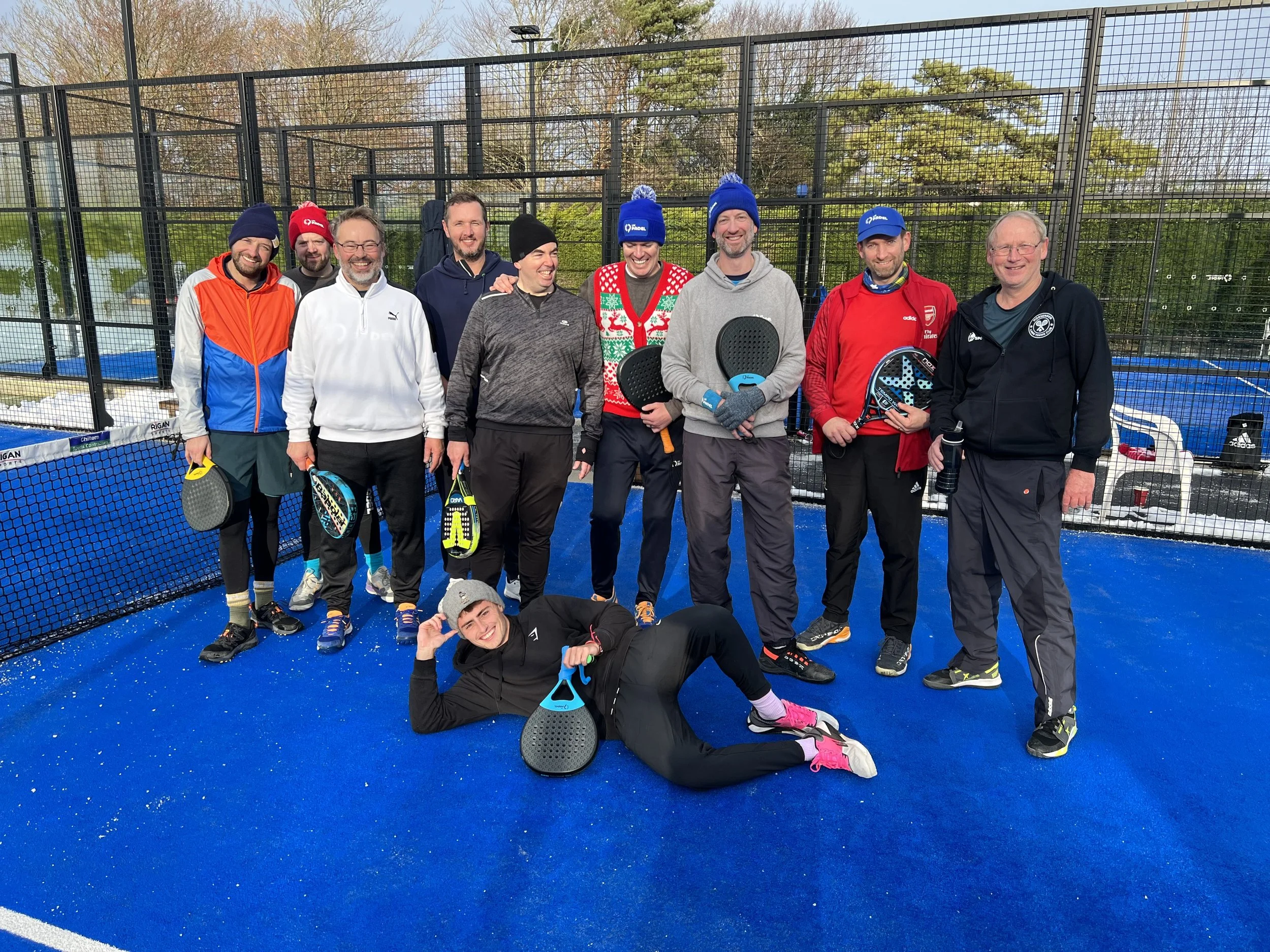 Group of ten people standing on a blue pickleball court, smiling at the camera, holding paddles, wearing casual sports attire and winter hats, with a chain-link fence and trees in the background.
