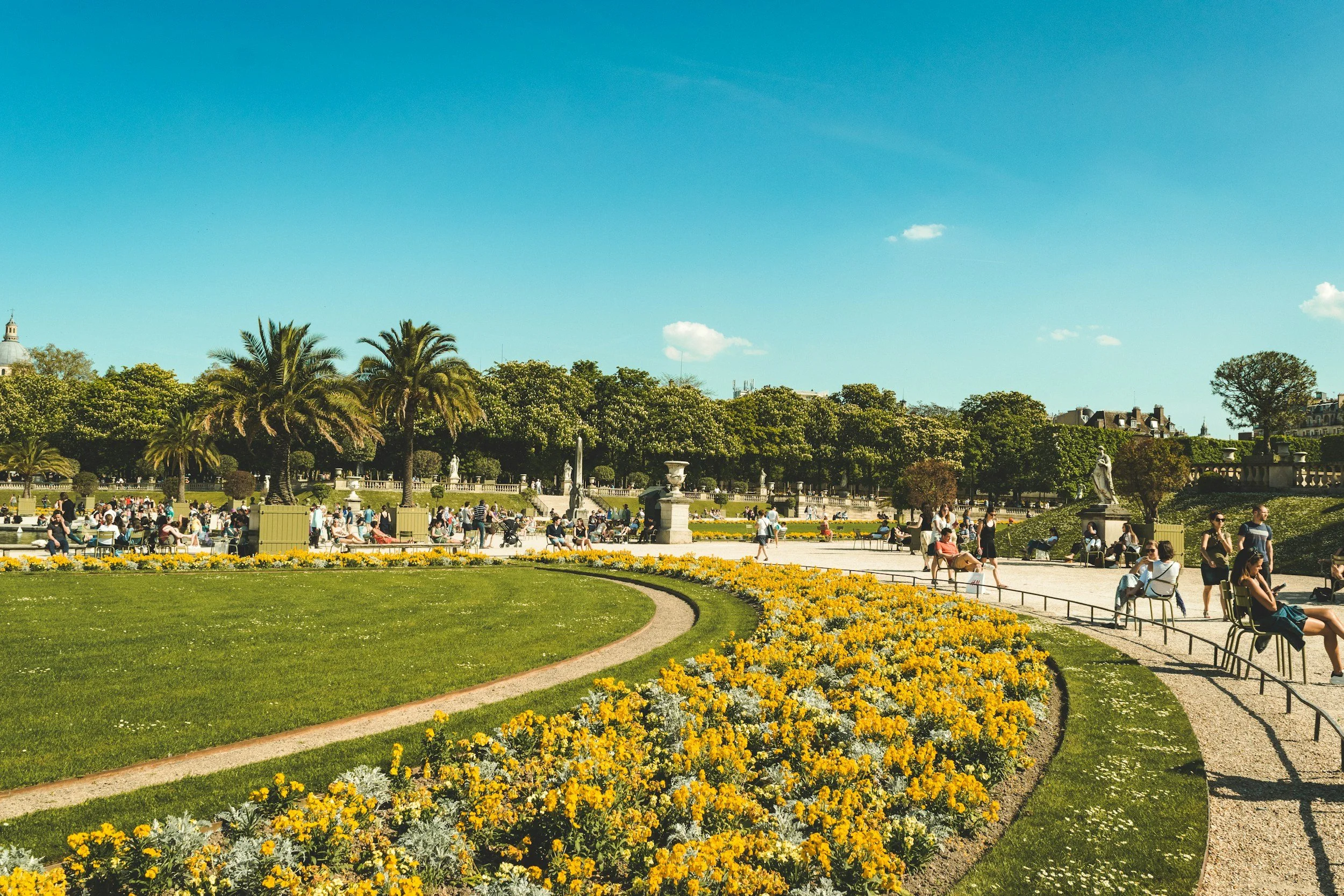 People walking and sitting in a large garden with yellow flowers, palm trees, statues, and a building in the background under a clear blue sky.