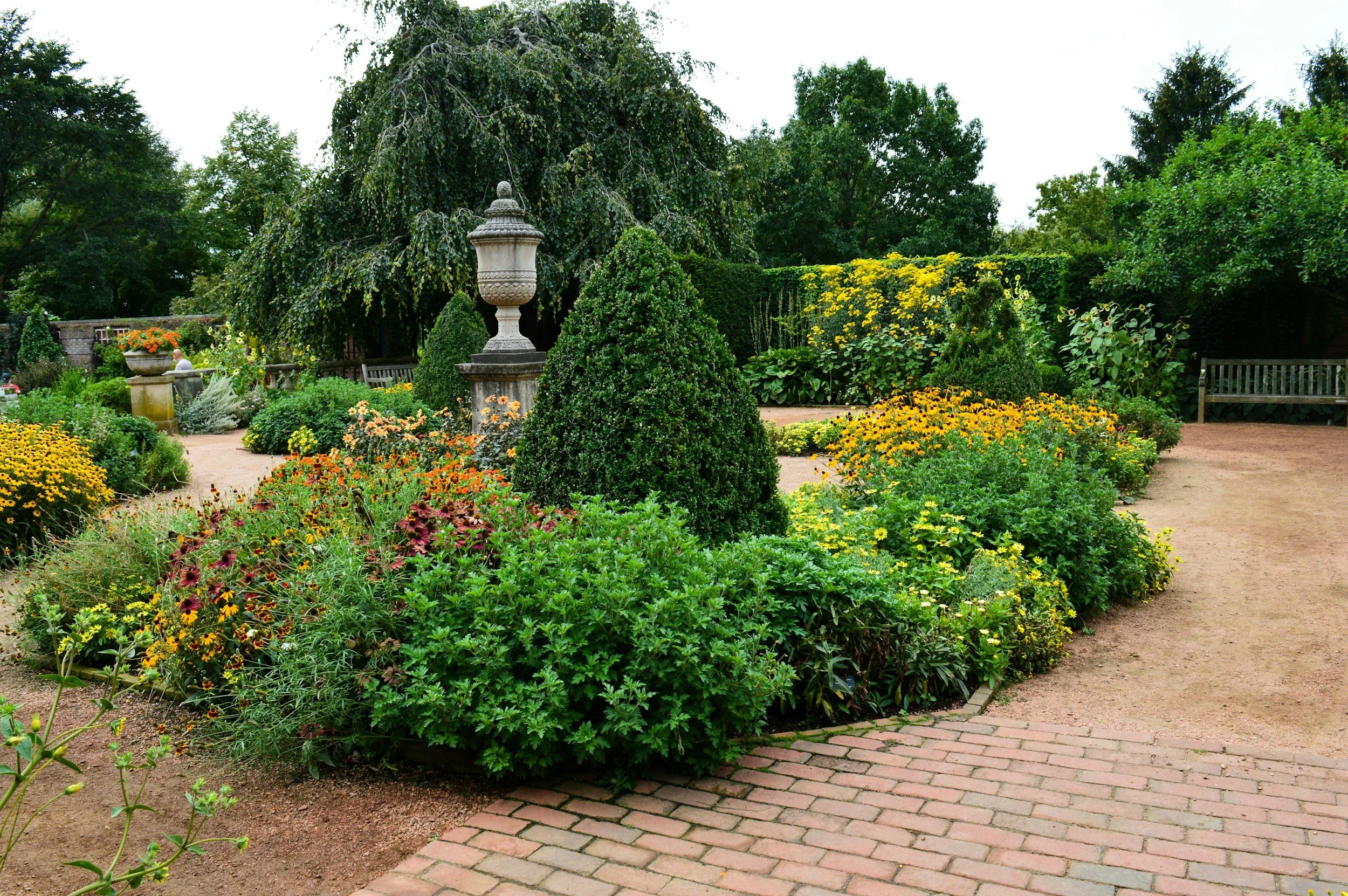 A lush garden with a variety of green plants, yellow flowers, and a large ornamental stone urn in the center. A paved brick path weaves through the garden, with benches and trees in the background.