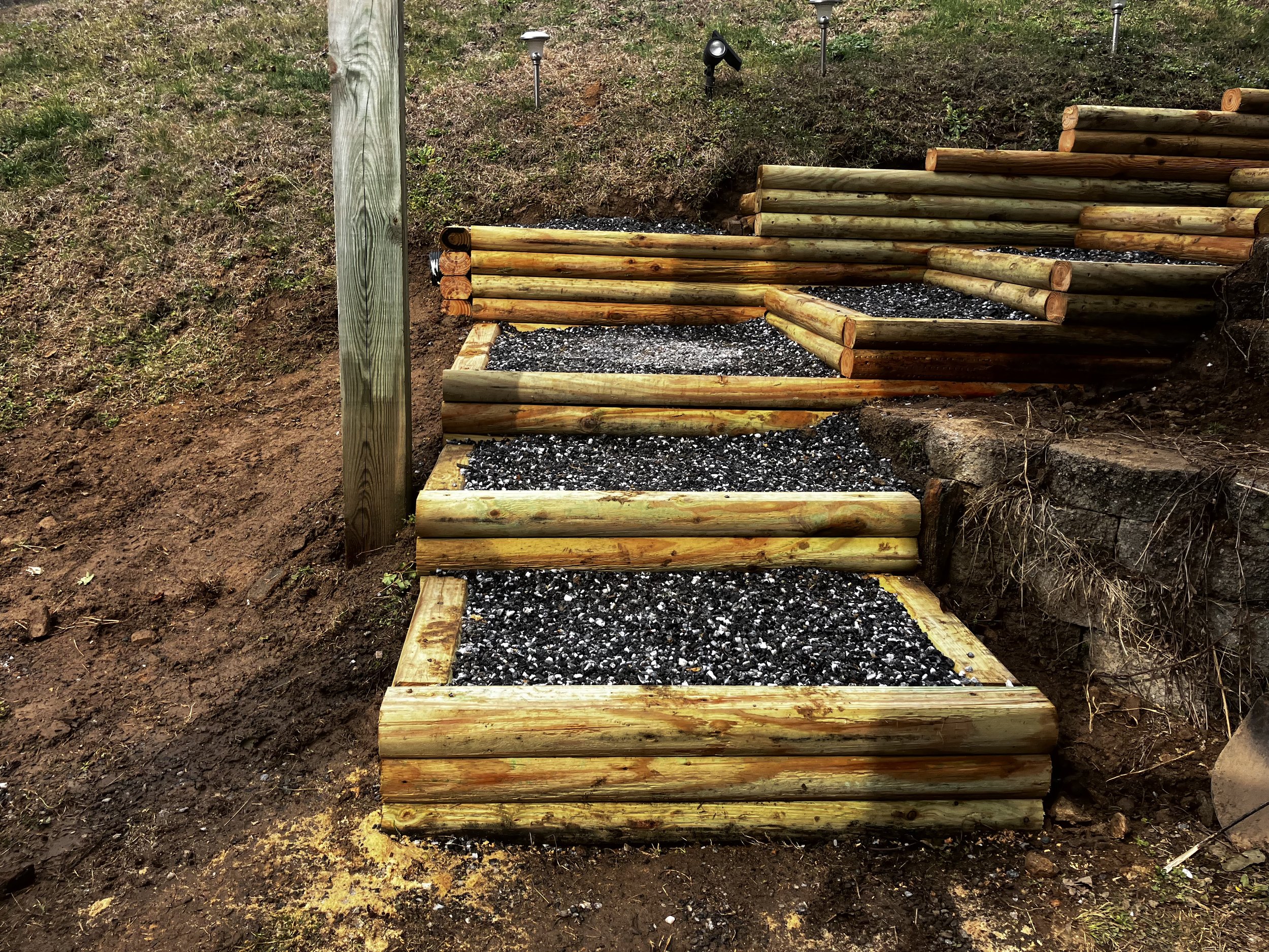 Wooden staircase under construction leading up a small hill, with black gravel on the stairs, surrounded by brown soil and grass on the hill.