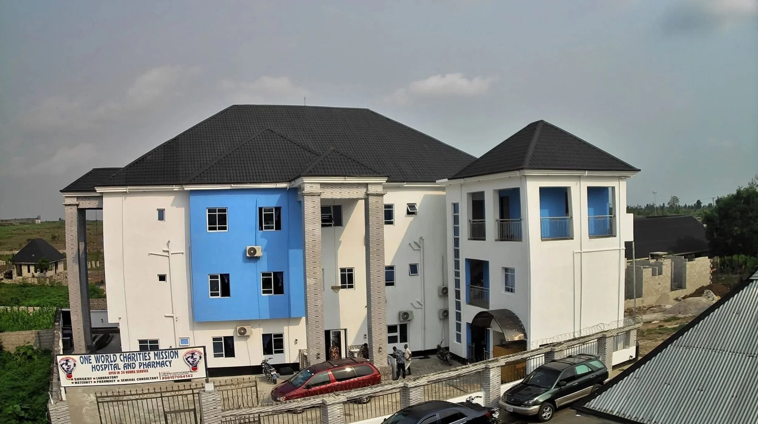 A multi-story building with white and blue exterior walls, multiple windows, and small balconies, located in a semi-rural area with a sign for a charity hospital and pharmacy in front, cars parked along the street, and cloudy sky overhead.