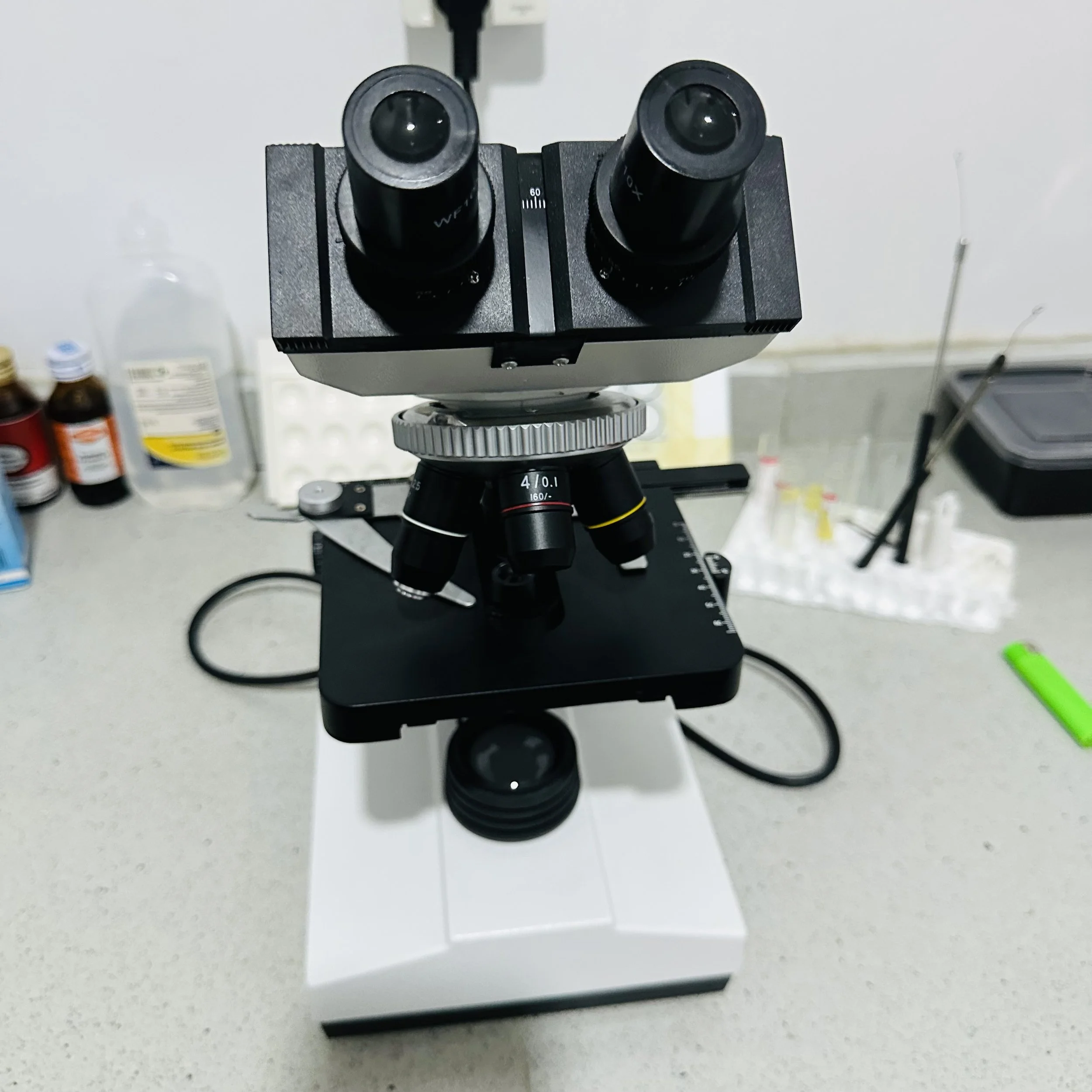 A microscope positioned on a lab table with bottles and supplies in the background.