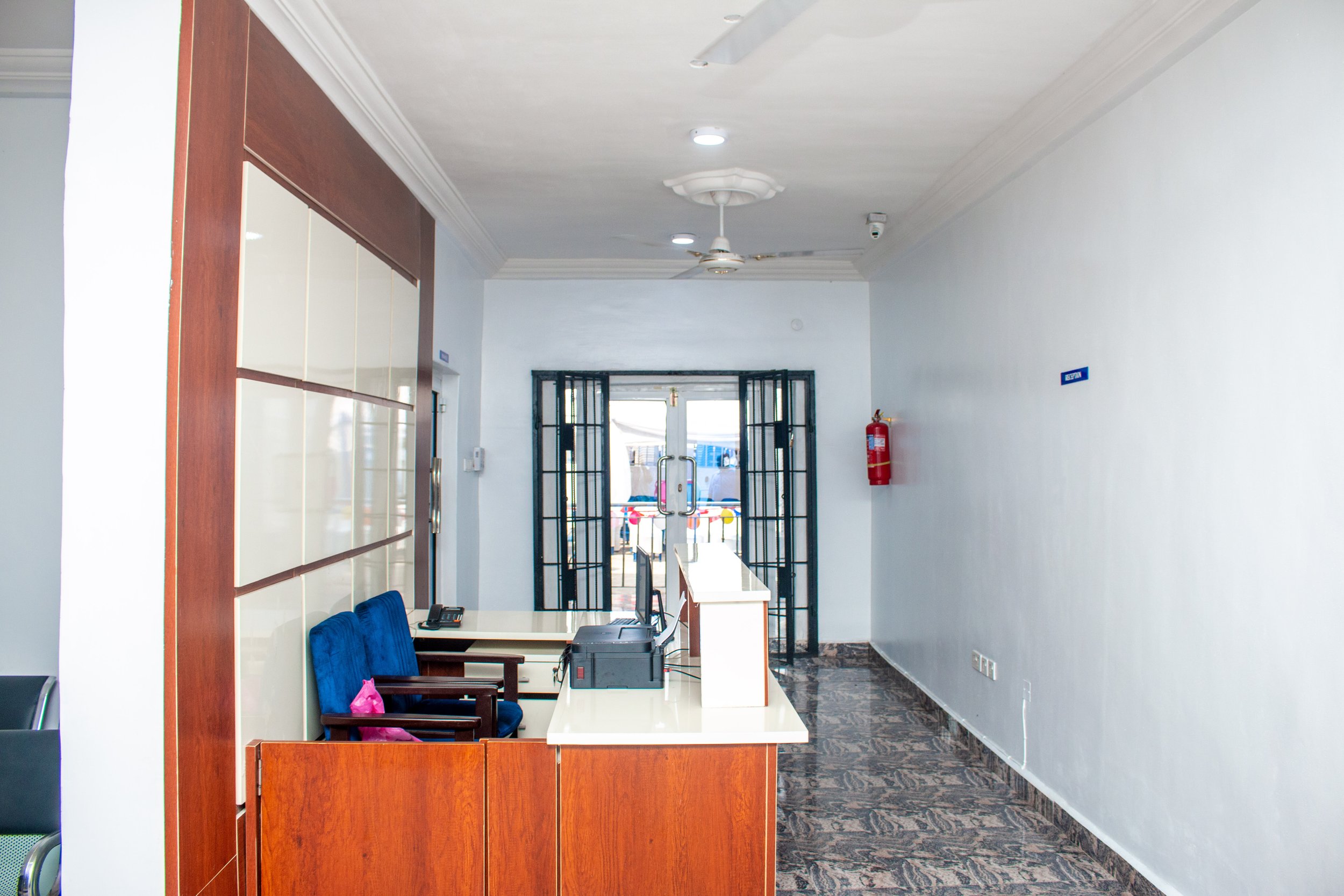 Interior of a reception or office area with a wooden counter, blue chairs, a computer, and a doorway leading outside.
