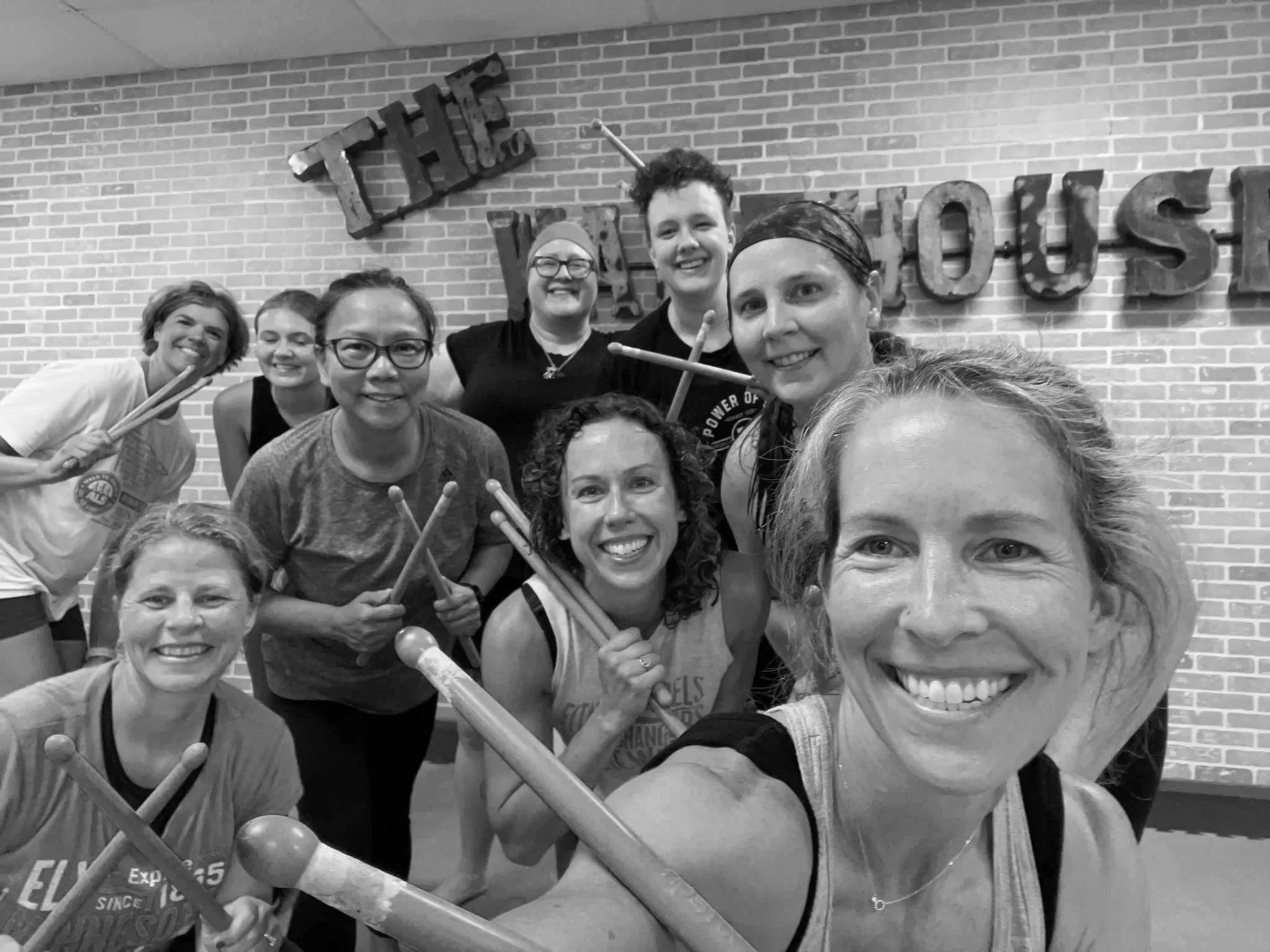 Group of diverse smiling women posing for a photo in front of a brick wall with large decorative letters and holding drumsticks.