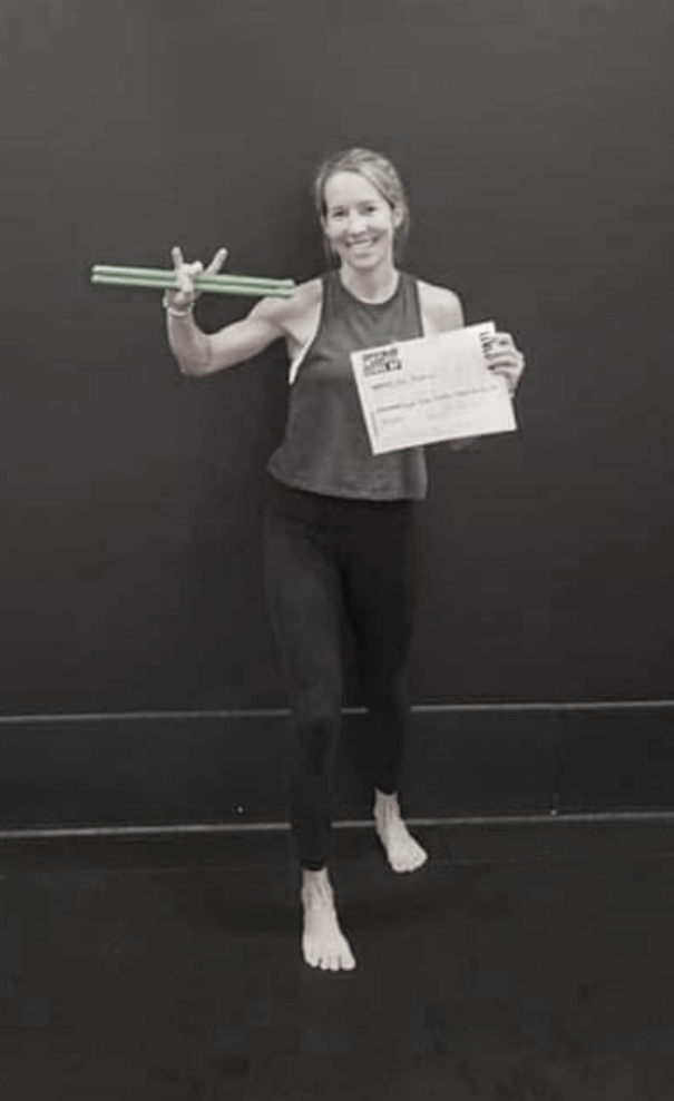 Smiling woman in workout clothes holding a certificate and a pair of green glow sticks, standing on a black floor against a dark wall.