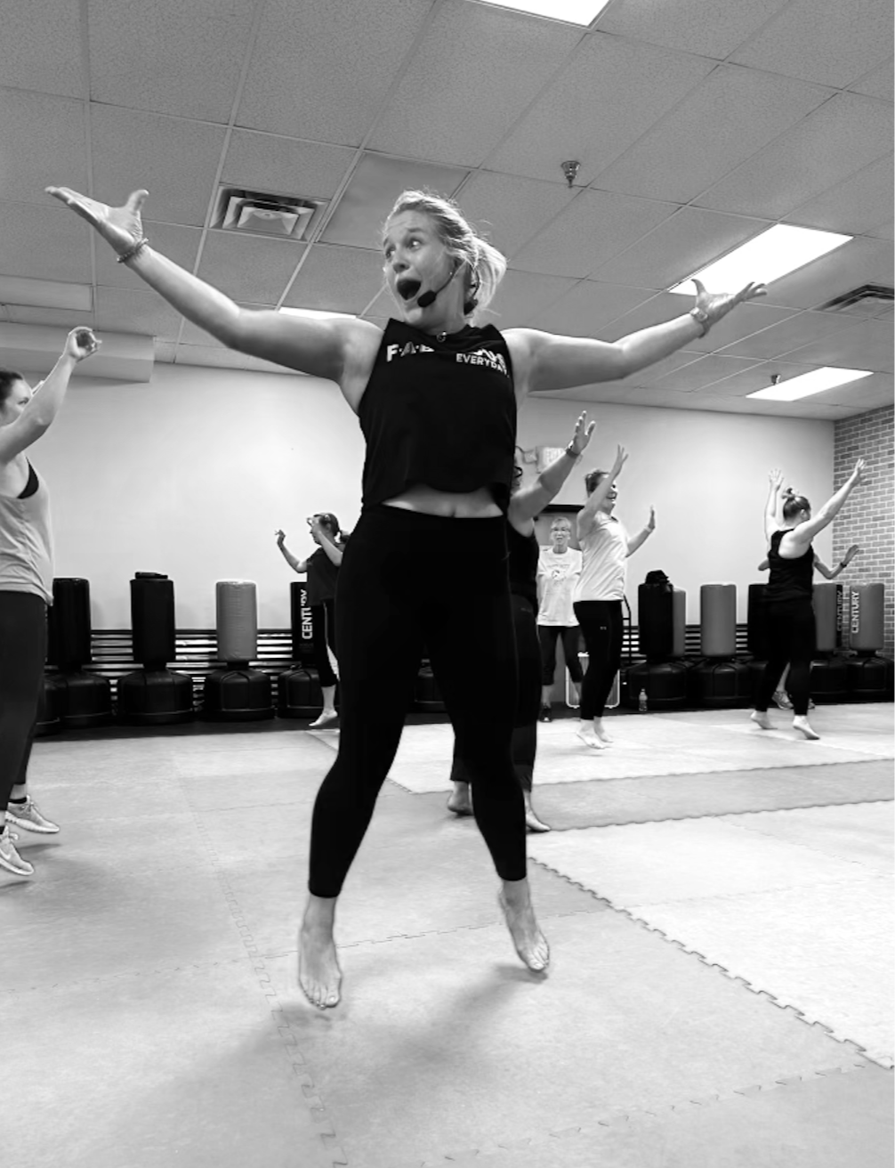 A woman with a headset demonstrating a fitness class in a gym, surrounded by other women participating in the workout.
