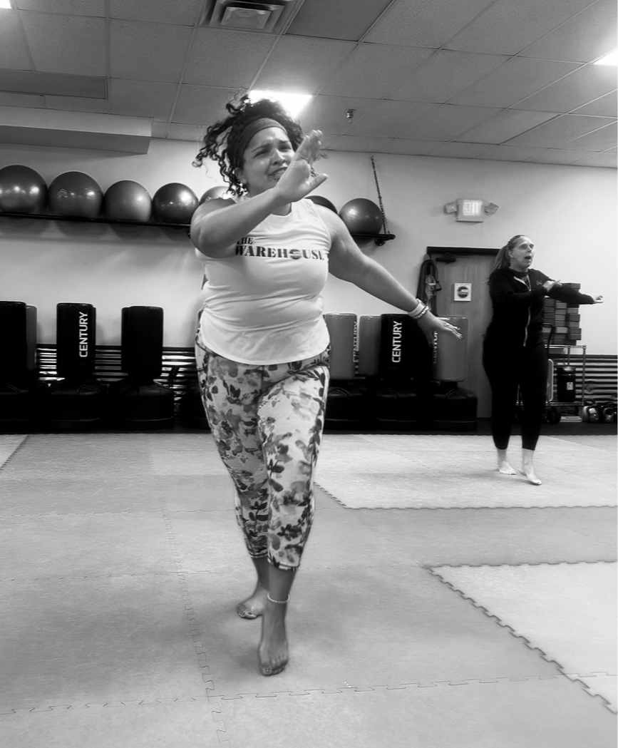 A woman exercising in a fitness studio with mats on the floor, some stability balls on a shelf, and punching bags along the wall. Another woman is also exercising in the background.