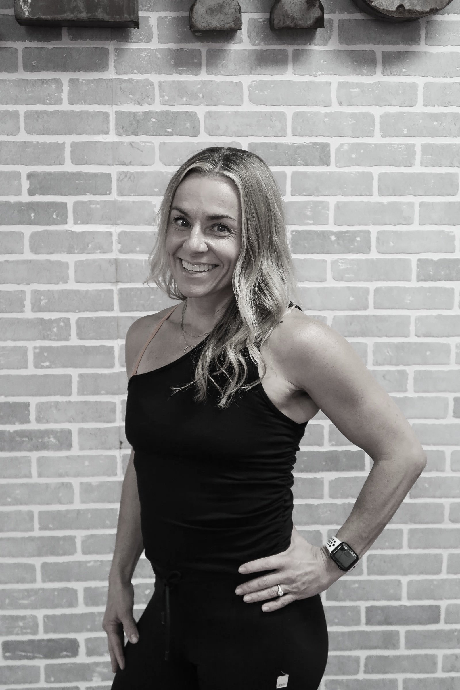 A woman with long wavy blonde hair is smiling, wearing a black sleeveless top, black pants, a wristwatch, and a ring, standing against a light-colored brick wall.