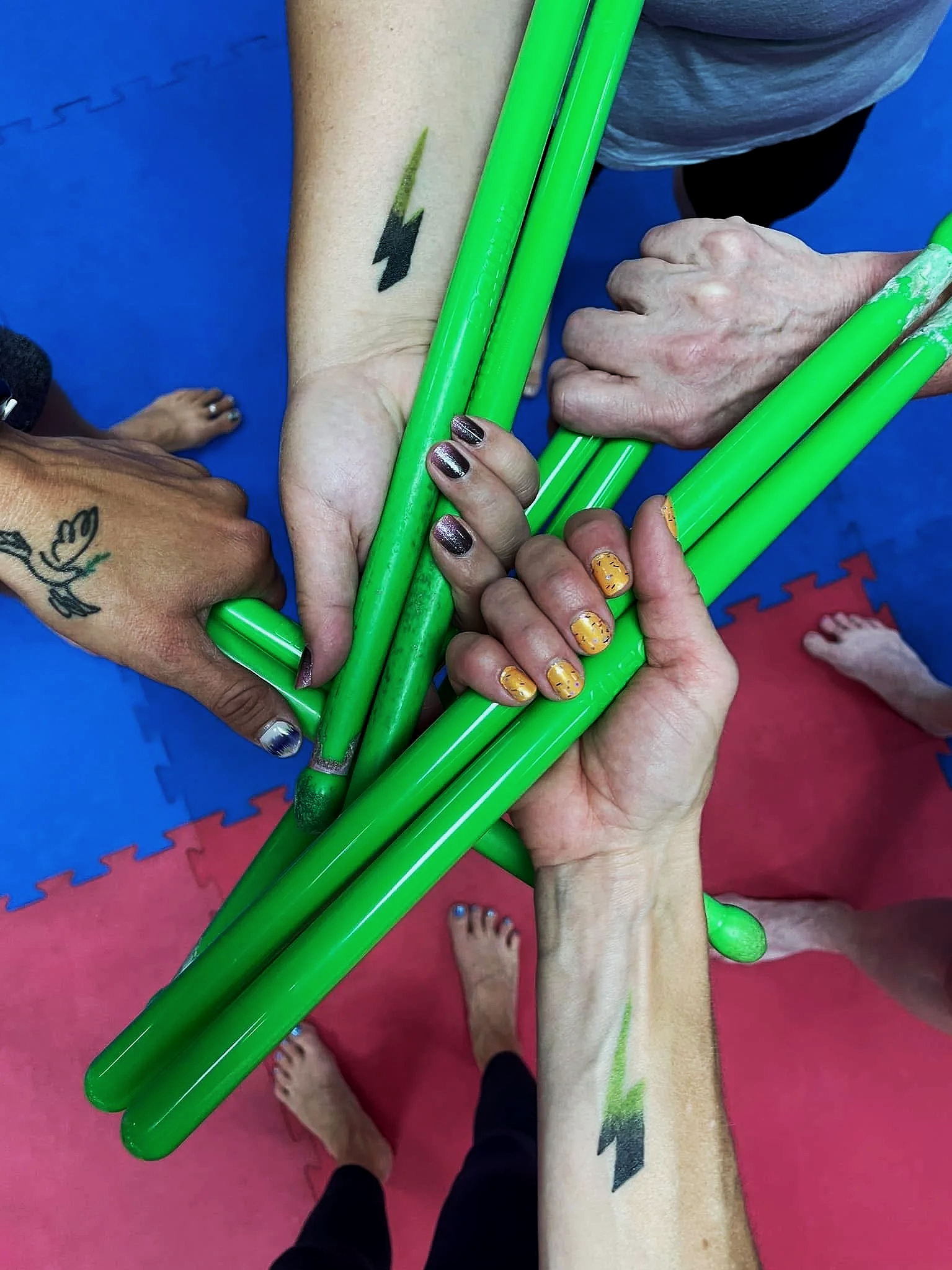 Top view of four hands gripping green plastic sticks together in a gym or martial arts setting, with a red and blue padded floor in the background and bare feet visible.