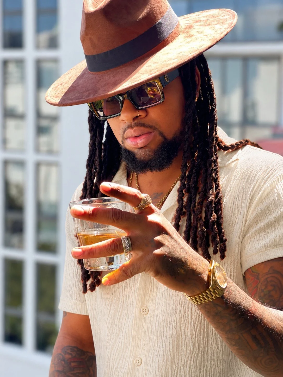 A man with dreadlocks wearing a wide-brimmed hat, sunglasses, gold watch, rings, and a light-colored shirt, holding a glass of whiskey outdoors.