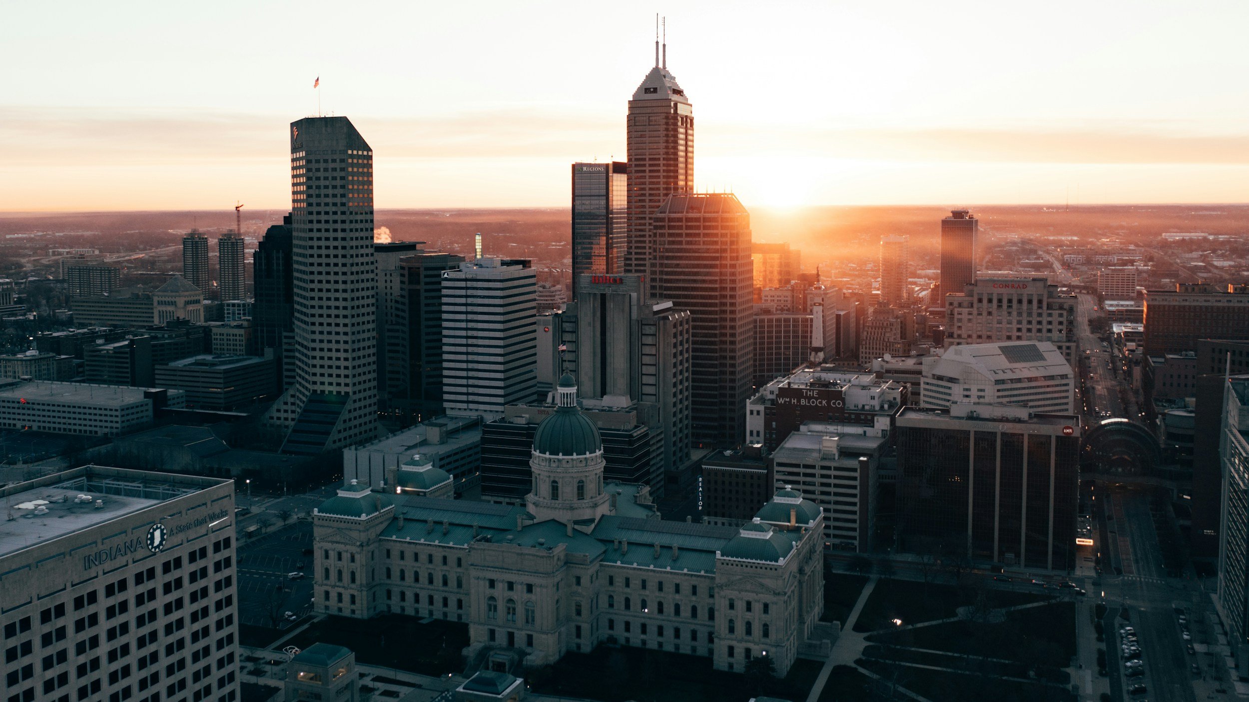 Sunset over a city skyline with tall skyscrapers and historic buildings, including the Indiana State House, in Indianapolis.