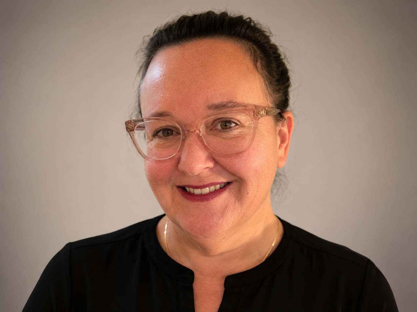 A woman with dark hair in a bun, wearing pink-framed glasses and a black top, smiling at the camera against a neutral background.