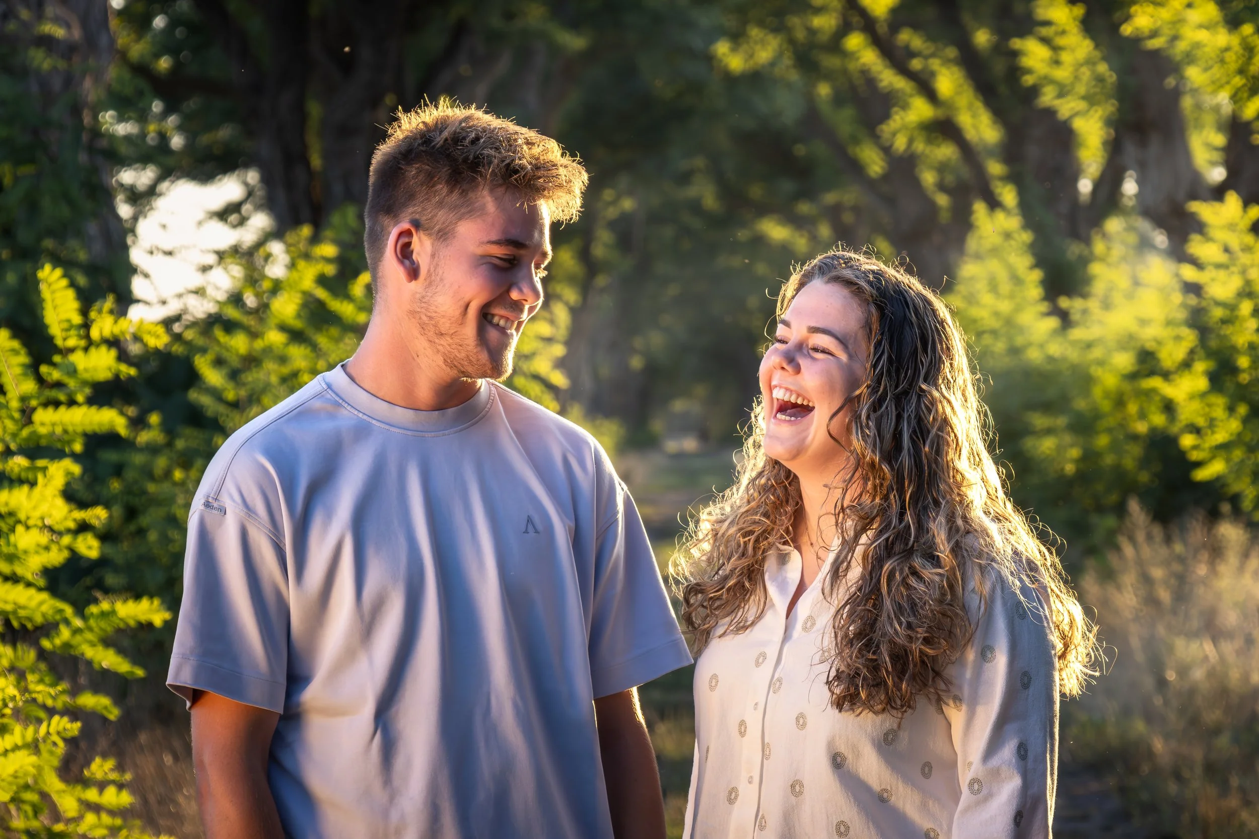 Twee jonge mensen, een man en een vrouw, lachen en praten samen buiten in een groene omgeving met zonlicht.
