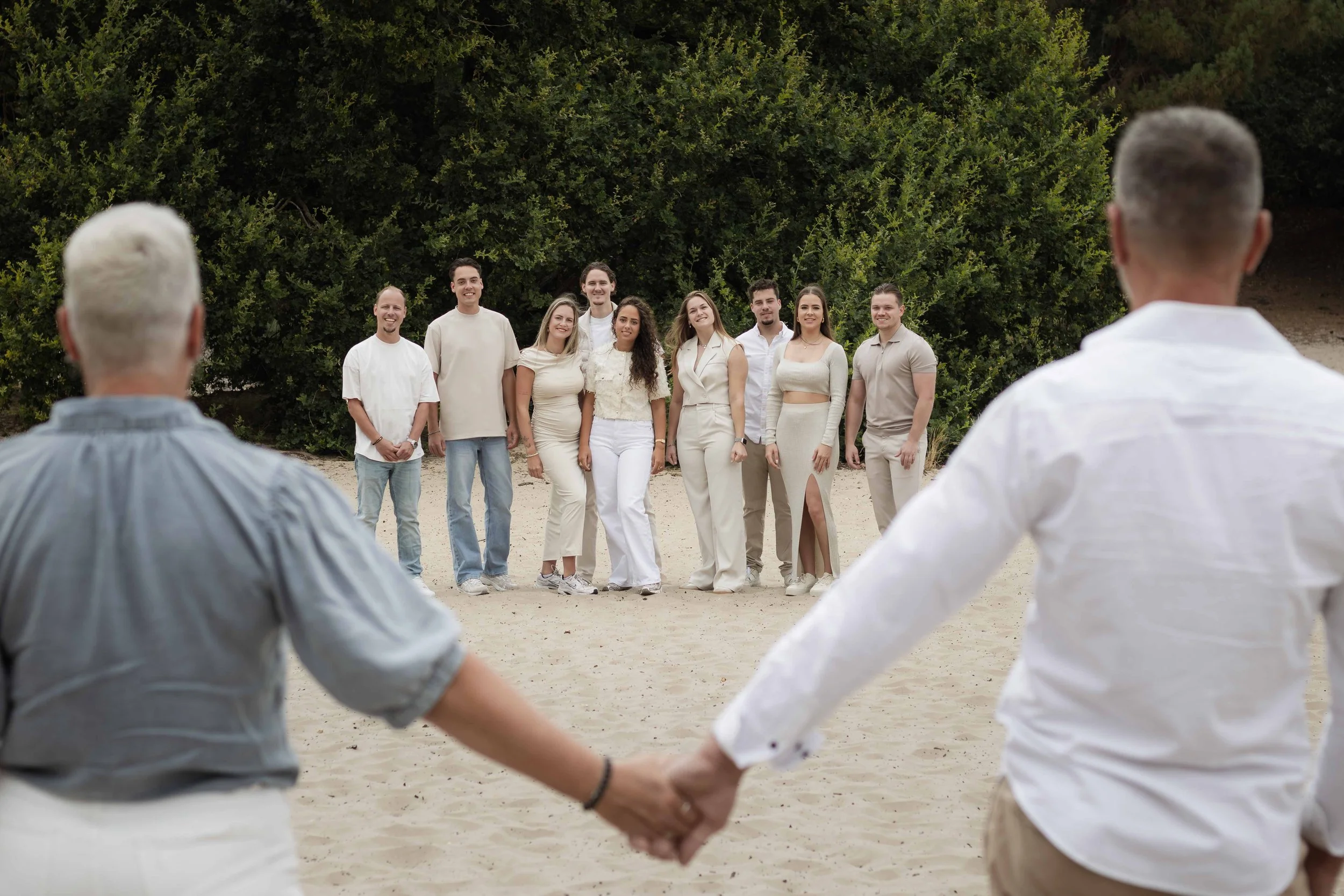 Groep mensen die hand in hand een kring vormen op het strand, met een groep mensen die voor hen staan en poseren voor de foto, terwijl groene bomen op de achtergrond te zien zijn.