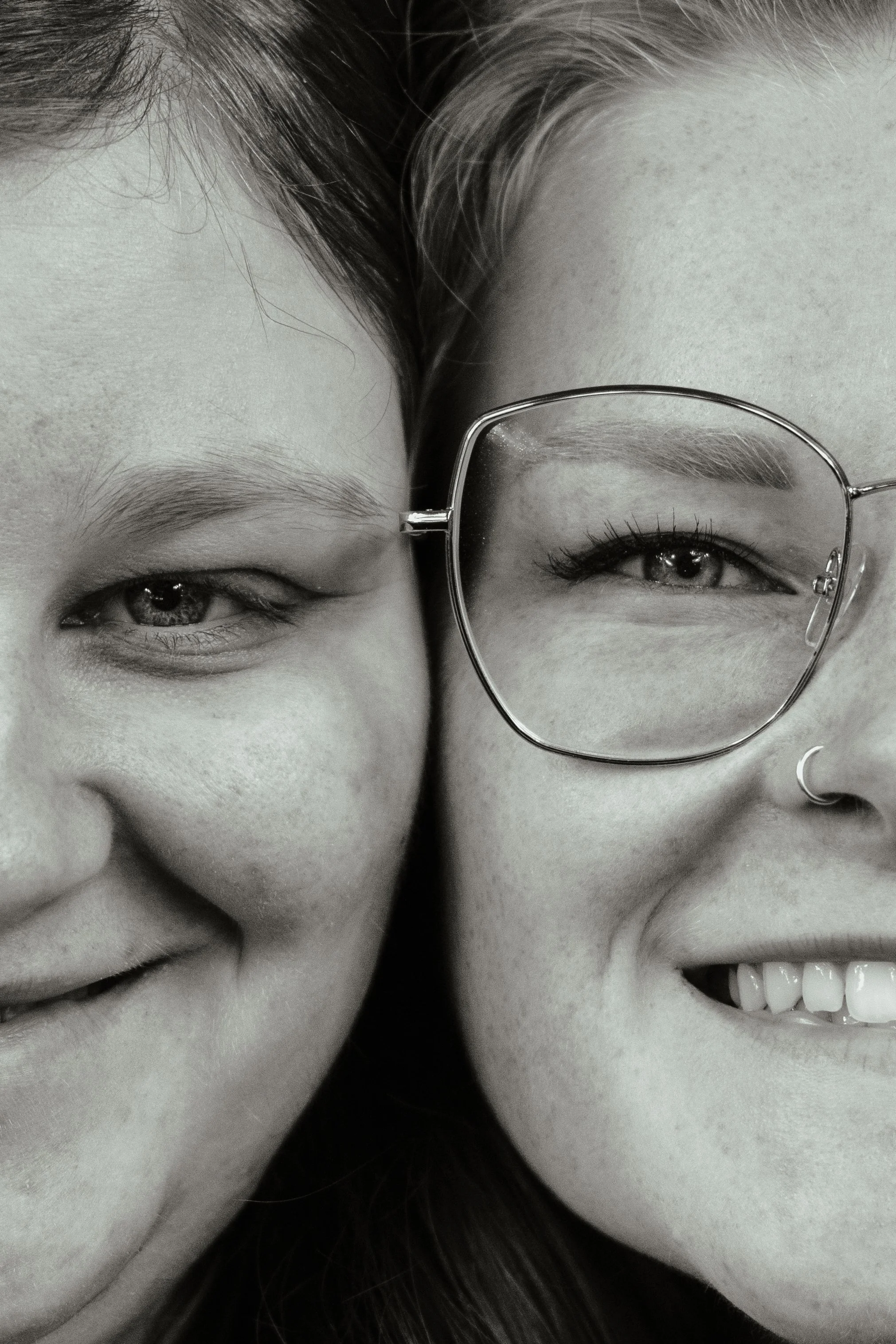 Close-up van twee mensen die dicht bij elkaar during een lach, een draagt een montuur bril, een glimlacht met zichtbaar gebit, in een zwart-wit foto.