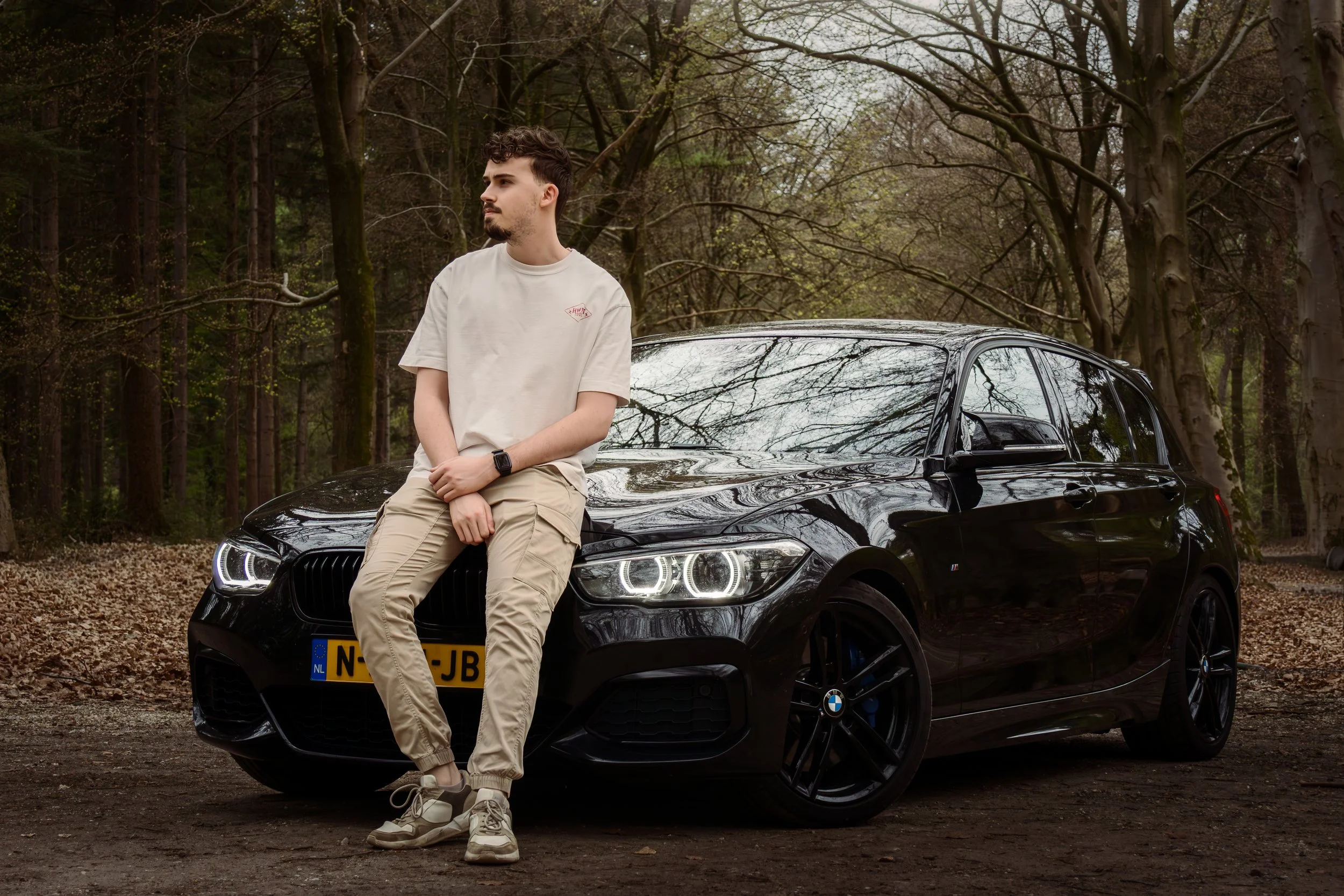 Man leunt tegen een zwarte BMW auto in een bosrijke omgeving met bladeren op de grond en bladeren aan de bomen.