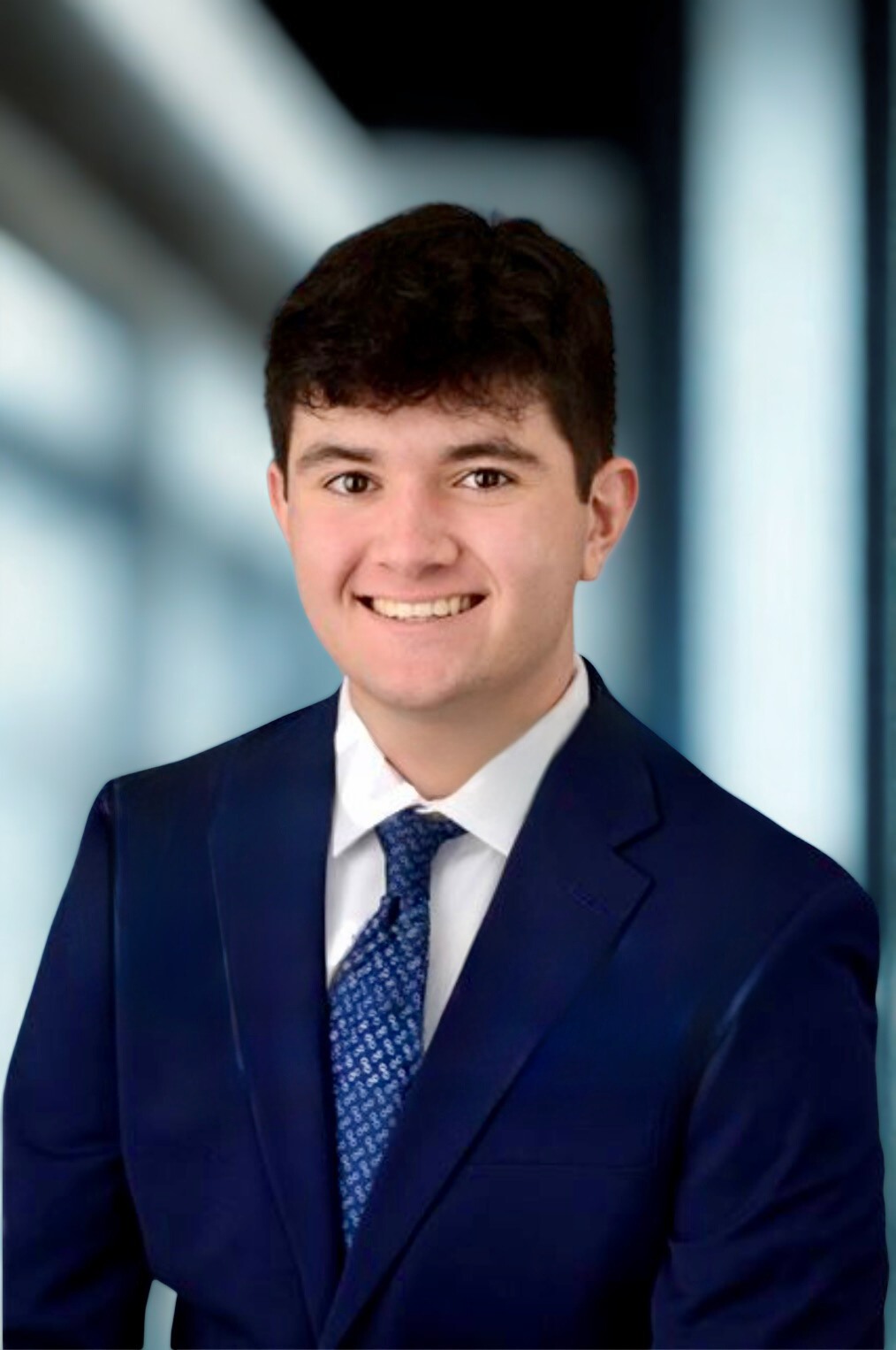 Young man in a navy suit with a white shirt and patterned blue tie, smiling, against a blurred blue and white indoor background.