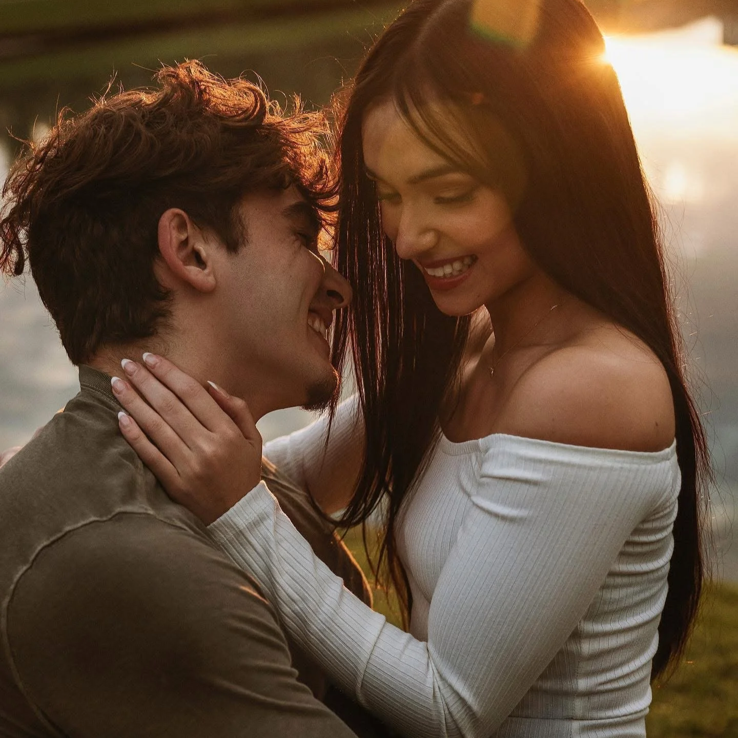 A young man and woman sharing a joyful moment outdoors at sunset, close together, with smiles and affection.