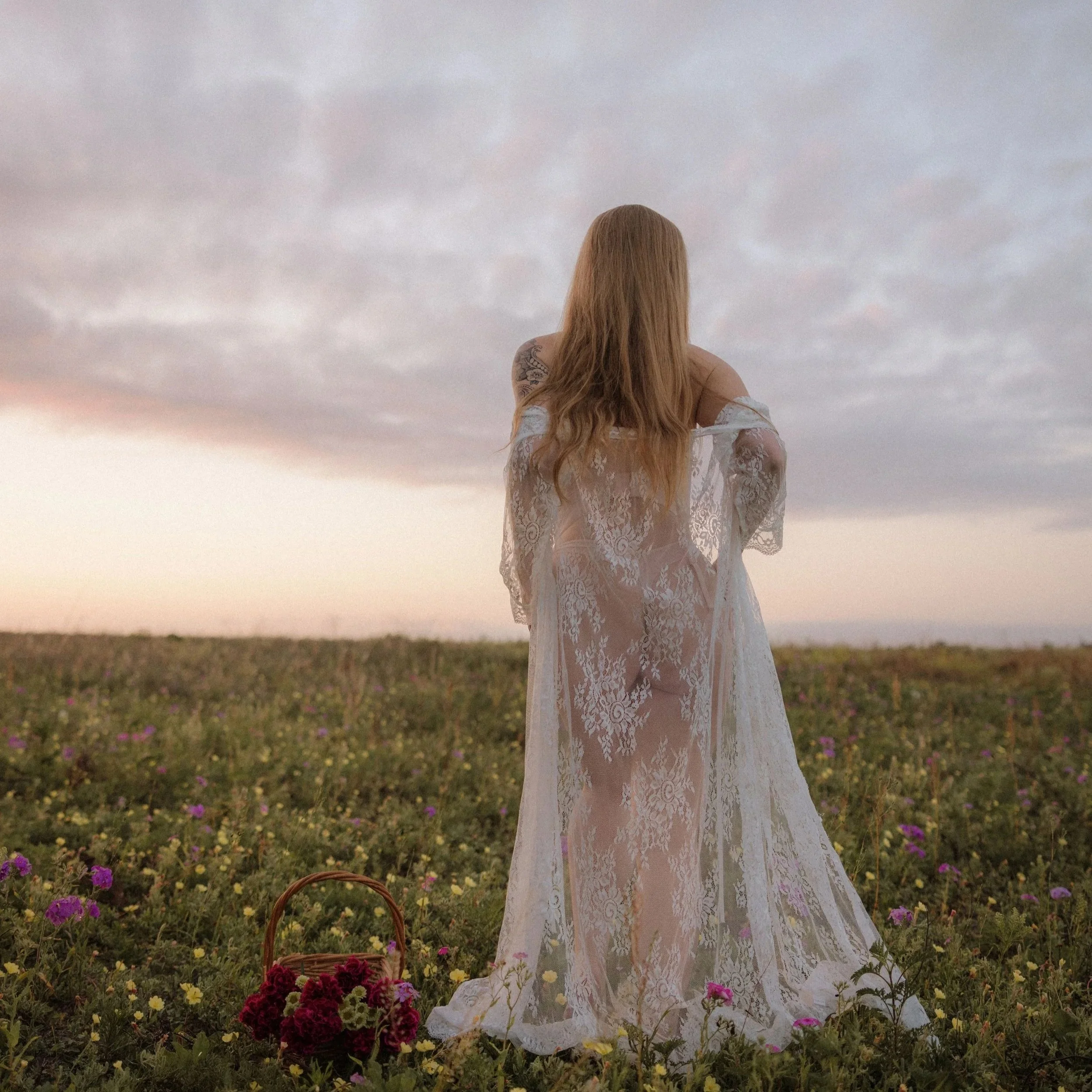 A woman in a white lace dress standing in a field of colorful flowers with a basket of flowers nearby, under a cloudy sky at sunset.